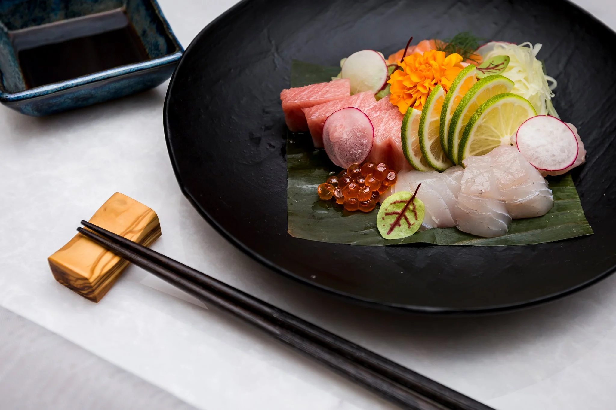 Sashimi platter with slices of raw fish, lemon and radish garnishes, orange fish roe, and edible flowers on a black plate with a payment terminal nearby.