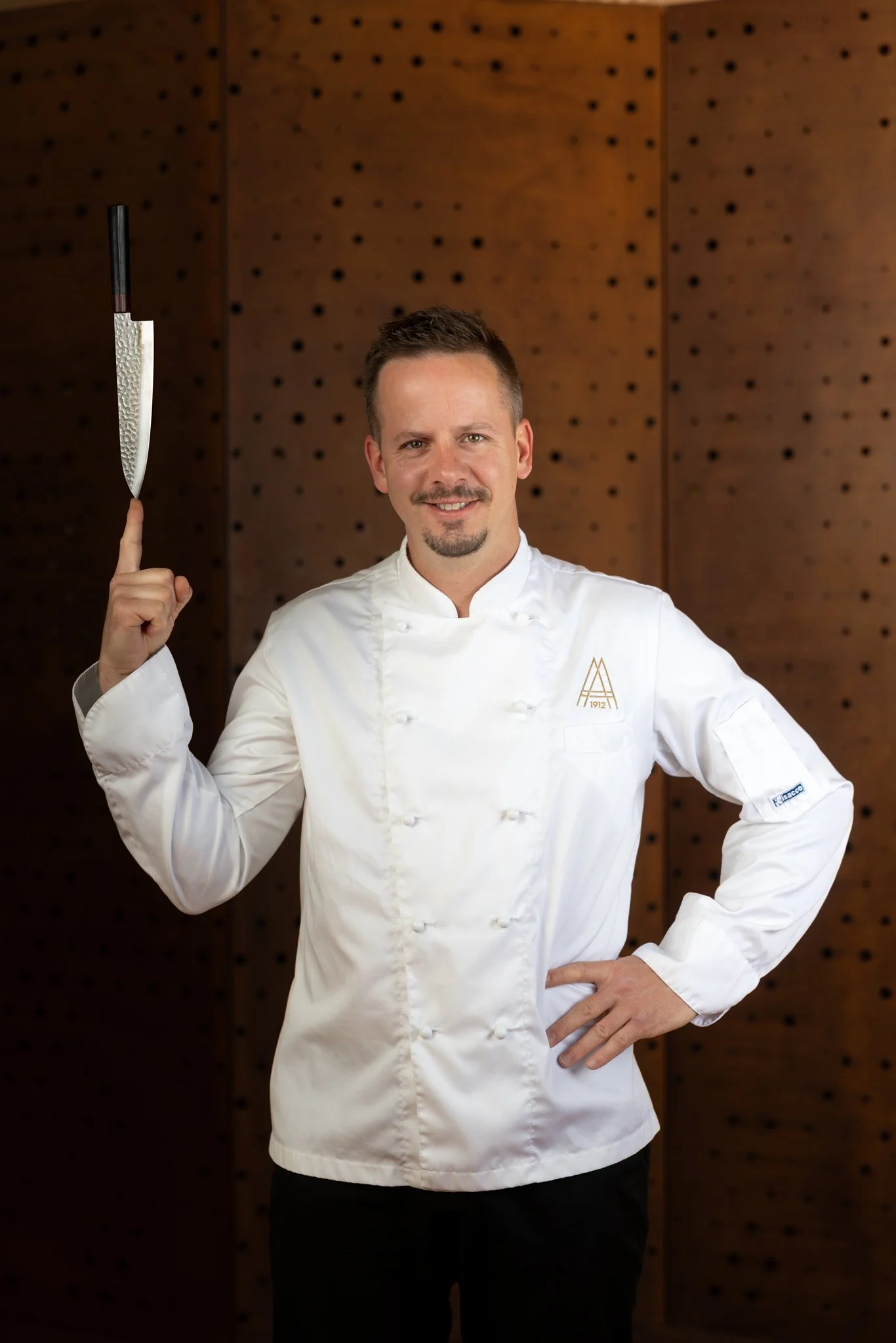 A male chef in a white chef coat smiling and holding a kitchen knife upright with his right hand, standing in front of a wooden wall with a pegboard pattern.