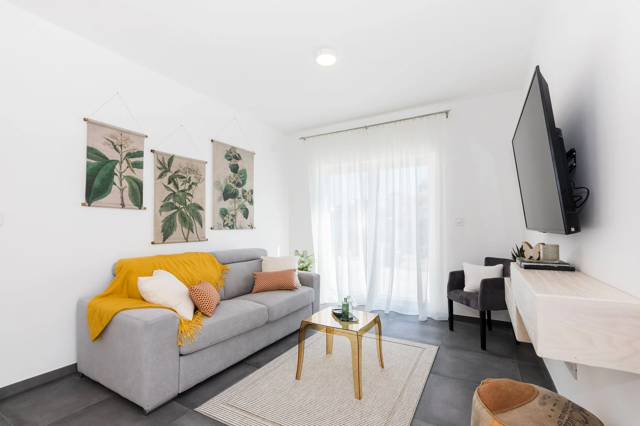 A living room with a light grey sofa draped with a yellow throw blanket and decorated with cushions, a small gold coffee table, botanical art wall hangings, sheer white curtains, a wall-mounted flat screen TV, a desk with items, and a small round foo