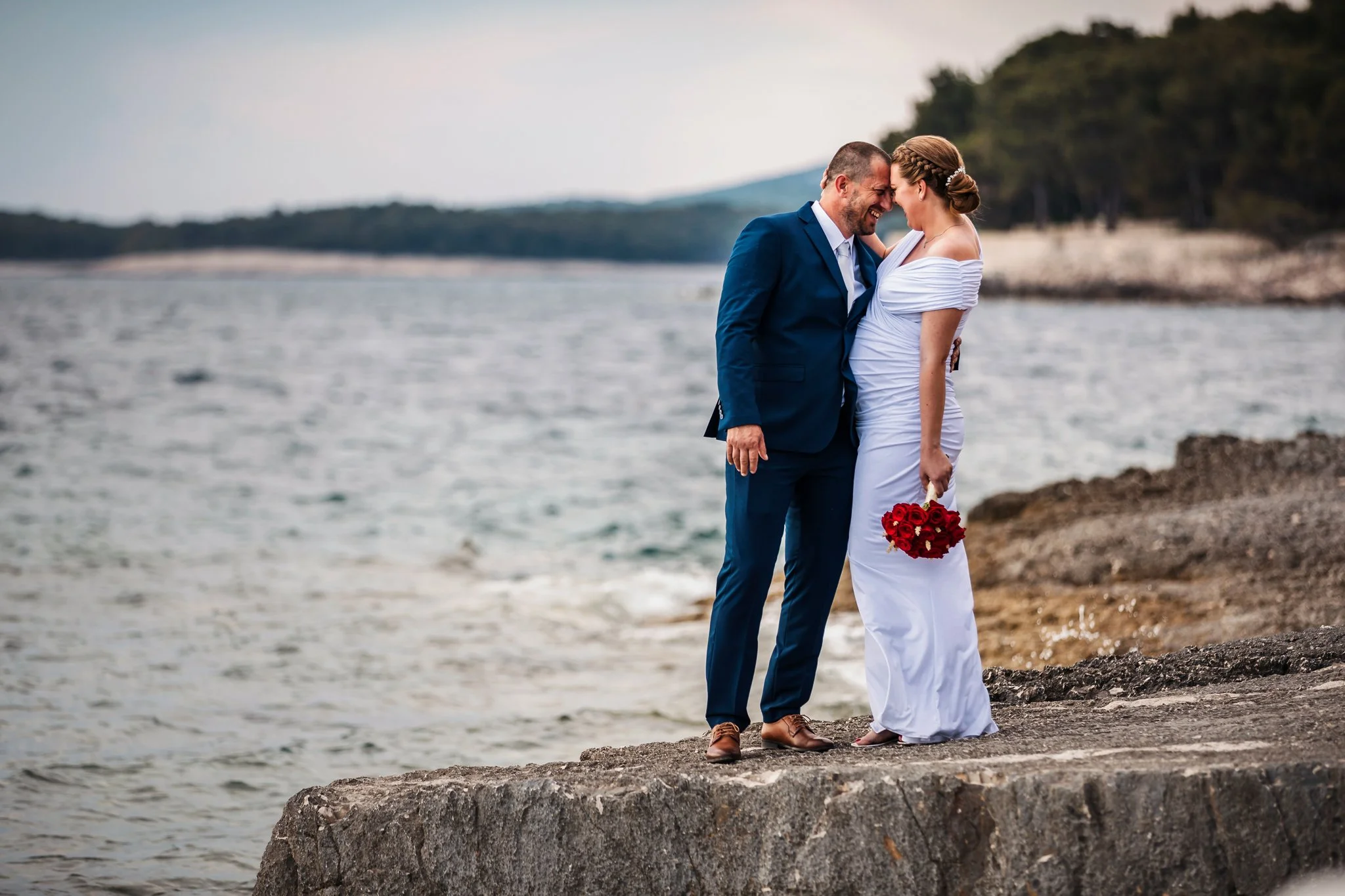 A couple in wedding attire standing on a rocky shoreline near water, smiling and touching foreheads, with the woman holding a bouquet of red roses.