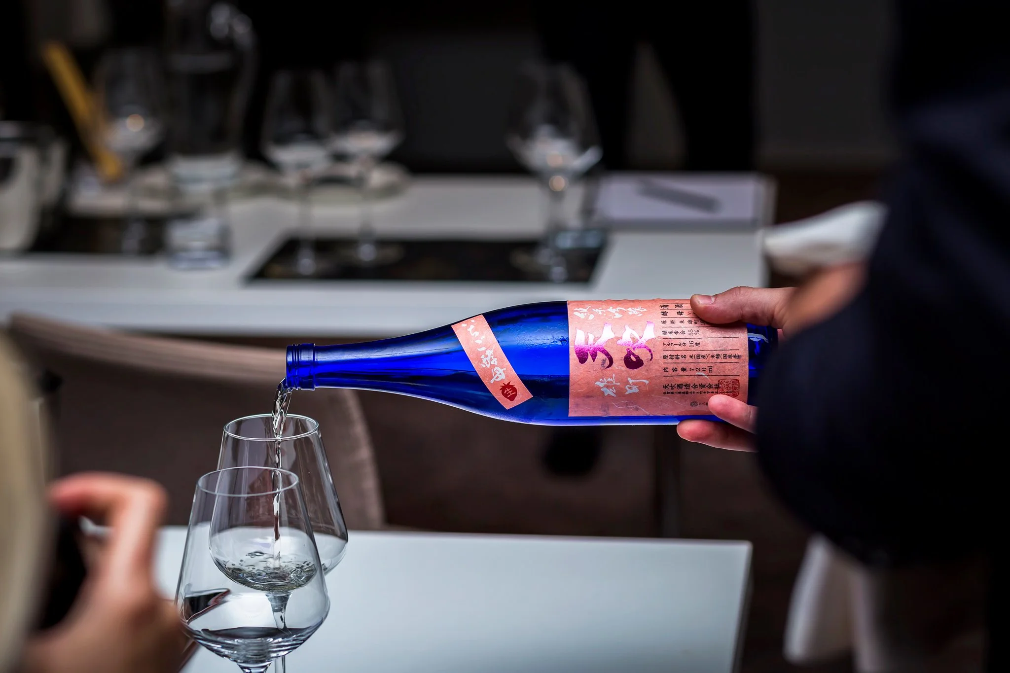 A person is pouring sake from a blue bottle into a wine glass, with two empty wine glasses on the table and a blurred serving area in the background.