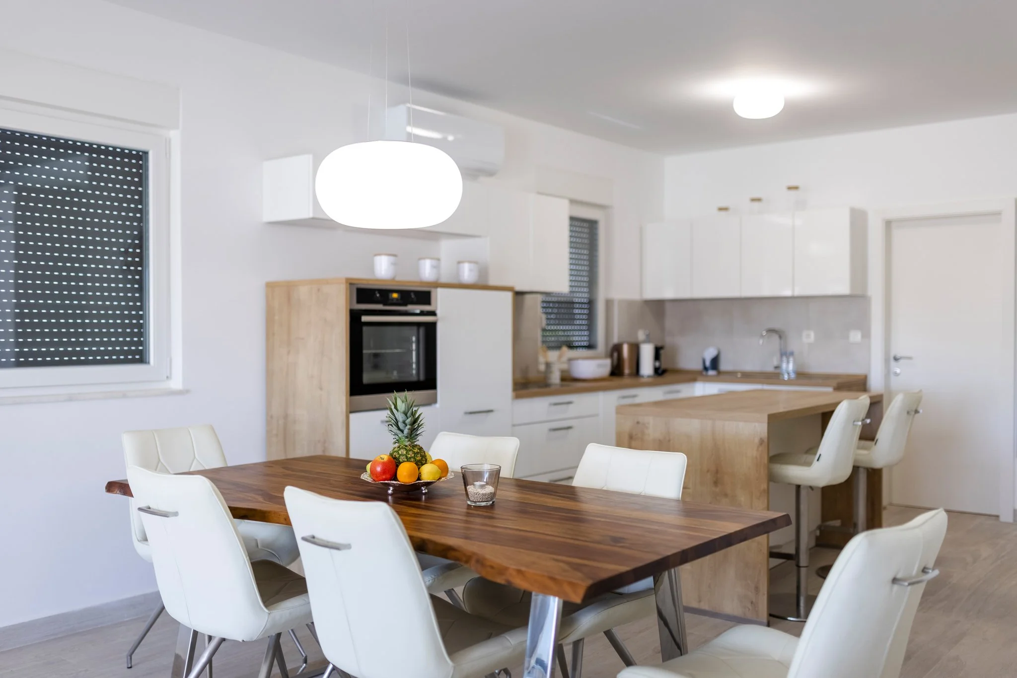 Modern kitchen with white cabinets, wooden countertops, and a wooden dining table with white chairs. A fruit bowl with pineapple, apples, and oranges is on the table. Bright lighting includes a ceiling light and a hanging pendant light. Windows with 