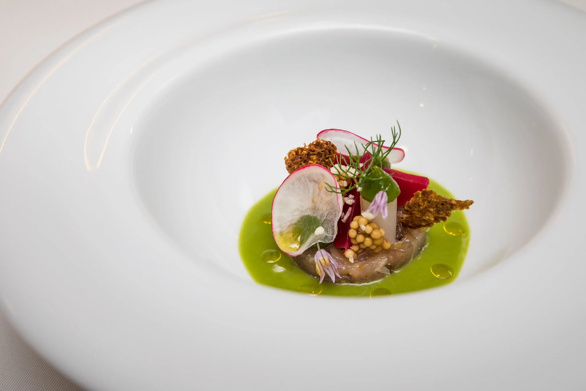 Elegant plated gourmet dish featuring thinly sliced radishes, microgreens, edible flowers, crispy garnish, and a vibrant green sauce on a white plate.