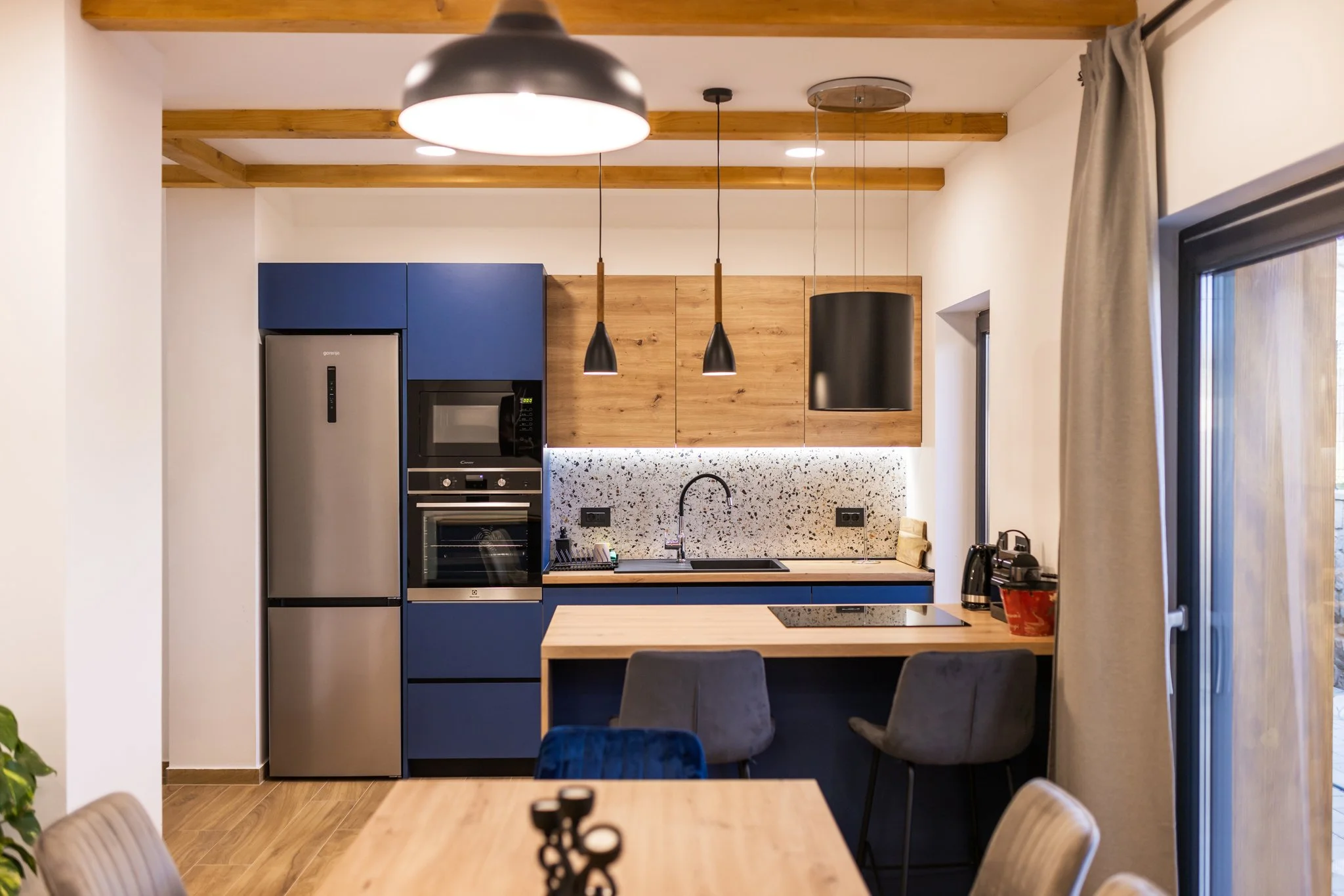 Modern kitchen with wooden ceiling beams, blue cabinets, black pendant lights, a black cylindrical hood, and a speckled white backsplash. There is a stainless steel refrigerator, microwave, and oven, with a coffee machine on the counter. Part of a di
