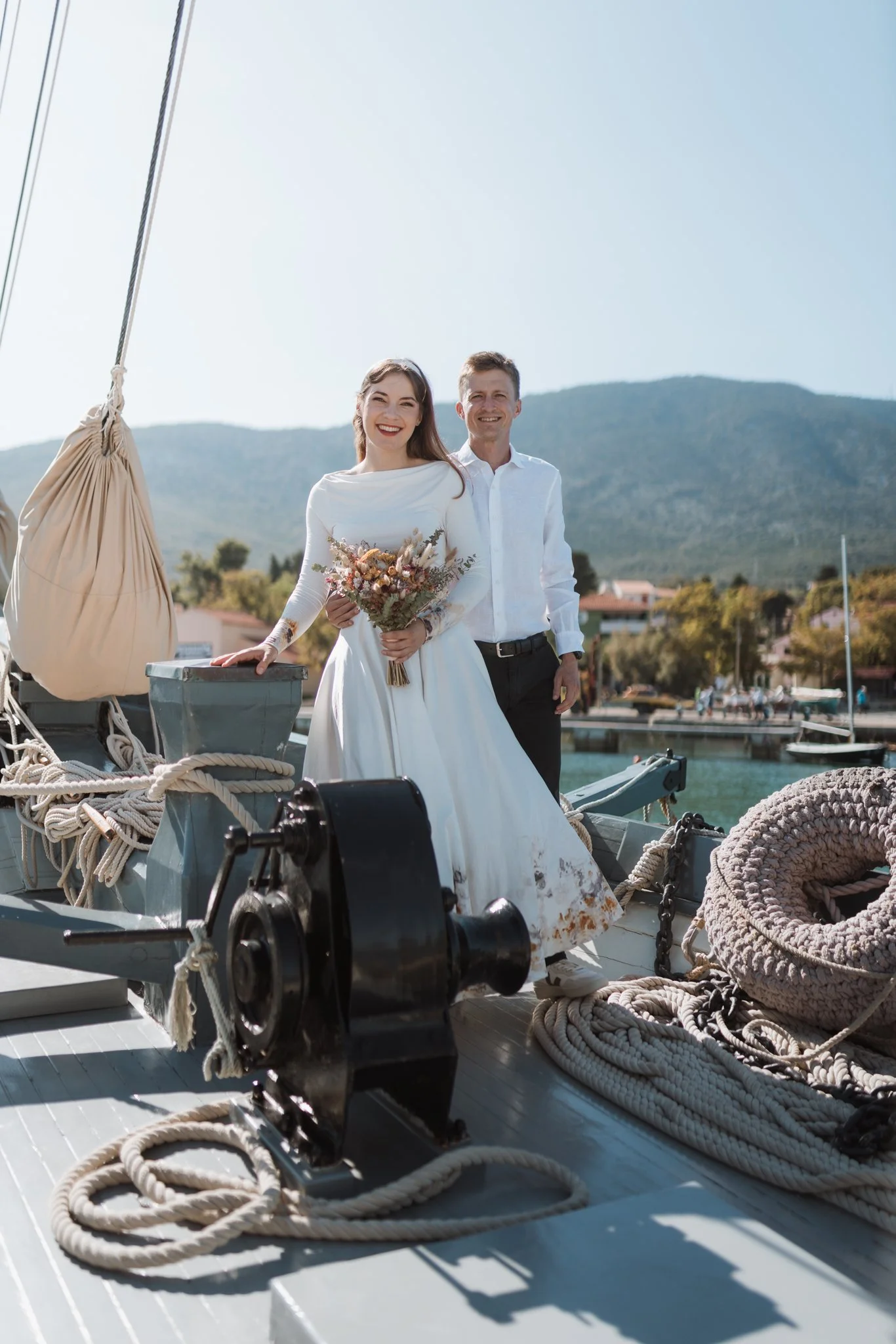 A smiling couple, woman in a white dress holding a bouquet and man in a white shirt, standing on a boat dock with ropes and nautical equipment, with mountains and houses in the background on a sunny day.