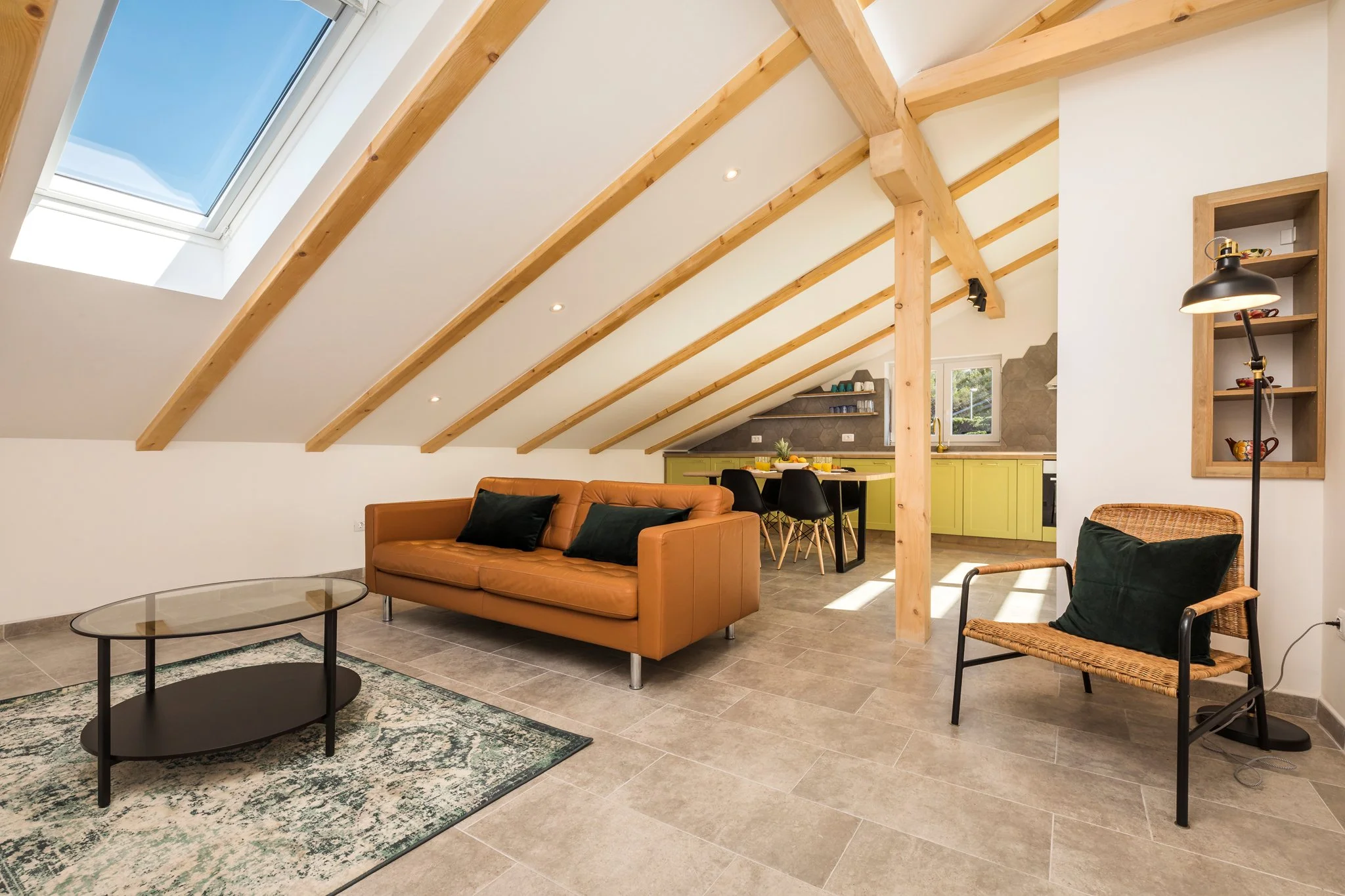 Living room with a tan leather sofa, black and wicker chairs, round coffee tables, skylight, wooden beams, open kitchen with lime green cabinets, and a decorative rug.
