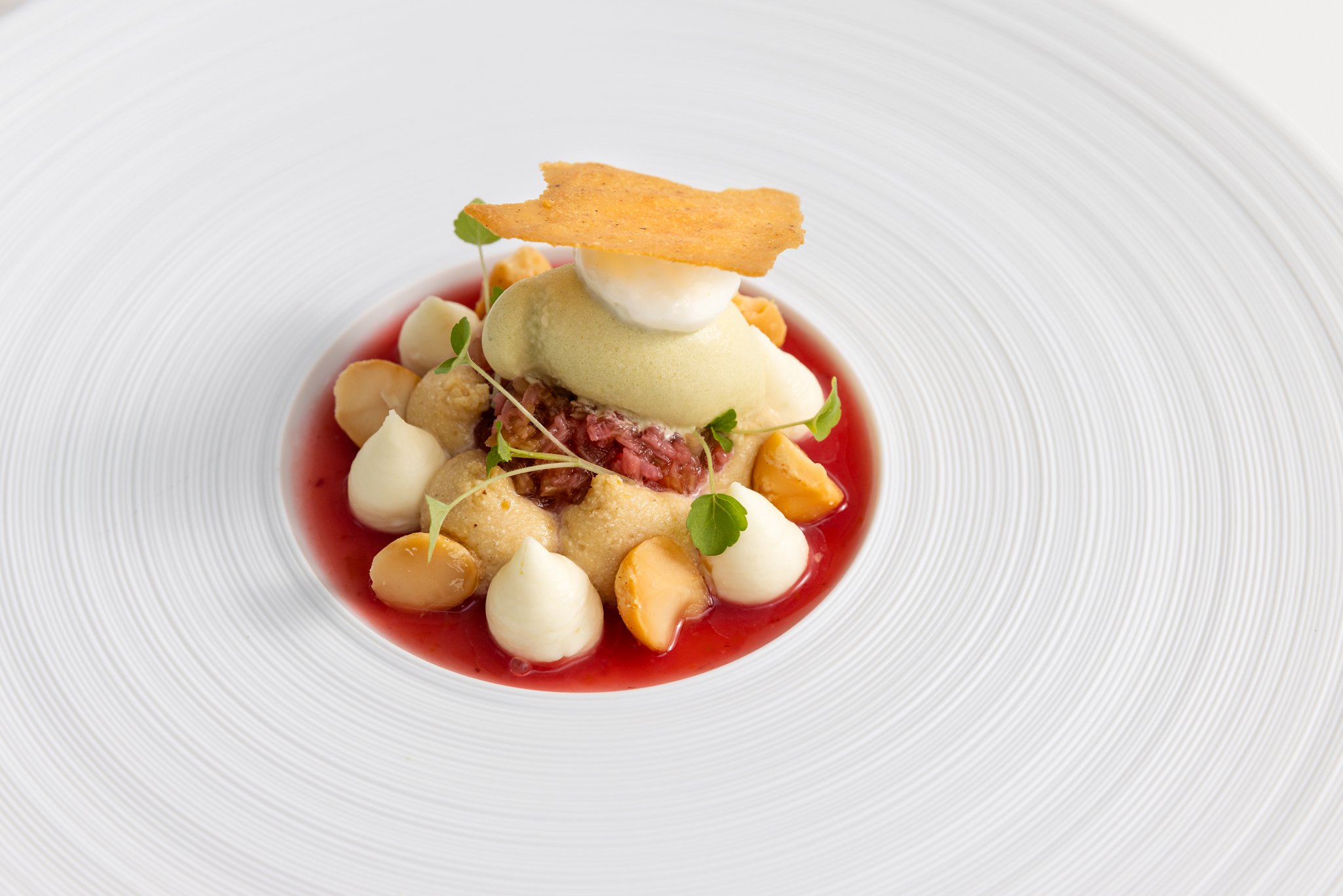 A plated dessert featuring a quenelle of pale green ice cream or mousse, topped with a thin crispy chip, surrounded by small dollops of cream, pieces of fruit, microgreens, and red sauce on a white textured plate.