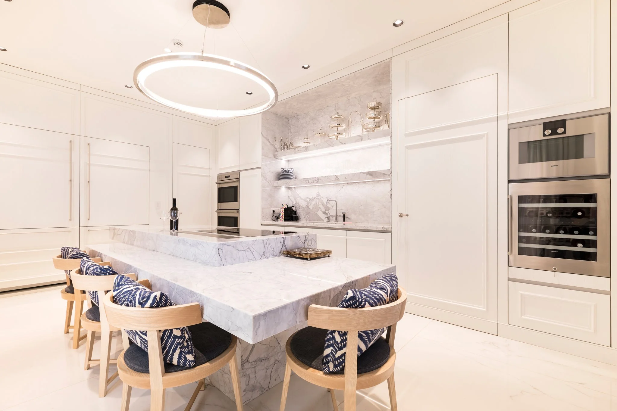 Modern kitchen with white cabinetry, marble countertops, and a marble backsplash. An oval-shaped hanging light fixture is above a large marble dining table with wooden chairs that have navy blue patterned cushions. There are double ovens and a wine r