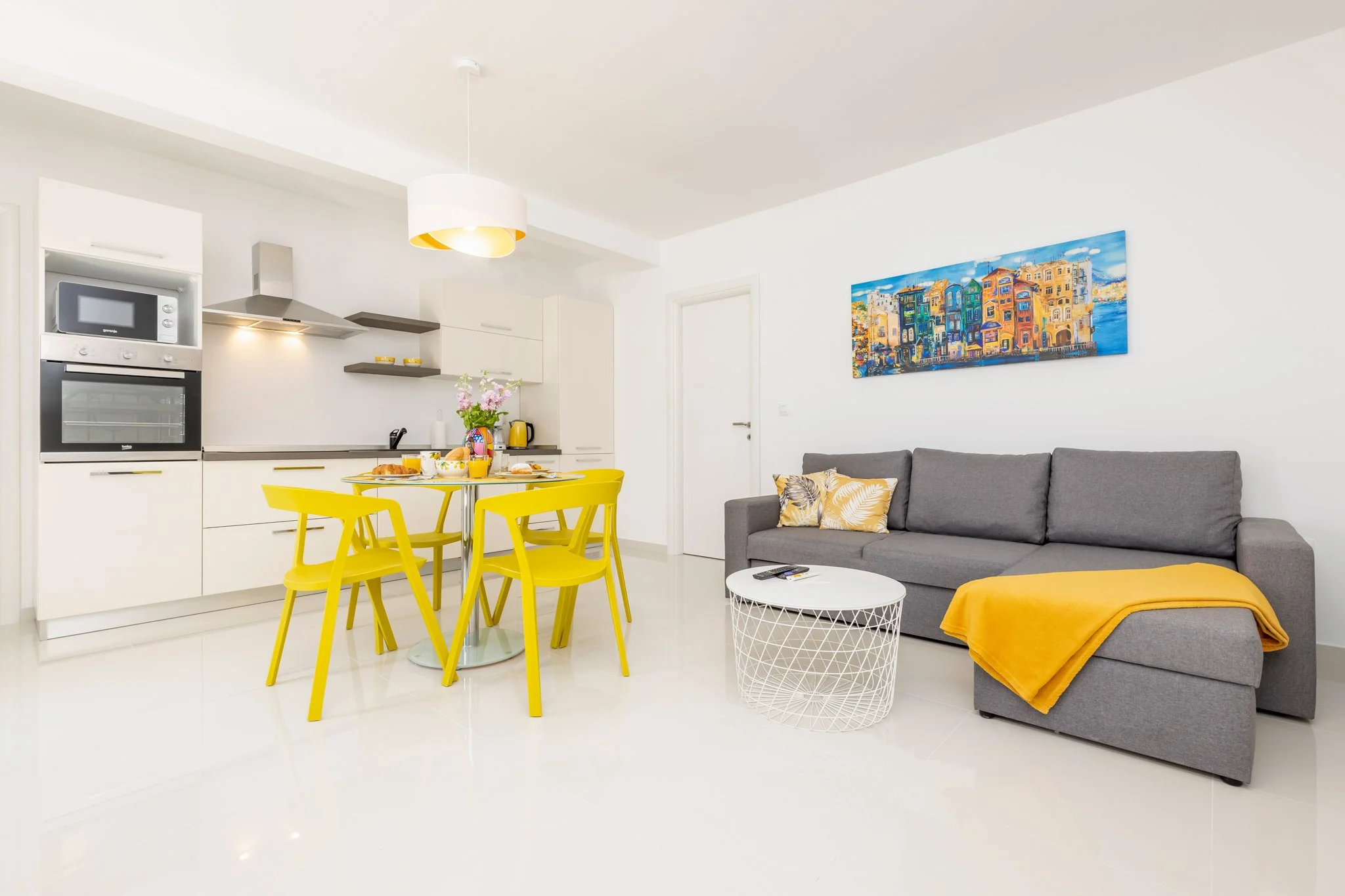 Bright, modern living space with an open-plan kitchen and dining area, featuring yellow chairs, white cabinets, and a gray sofa with yellow and leafy patterned pillows, a white round coffee table, wall art, and a white ceiling with a hanging light fi