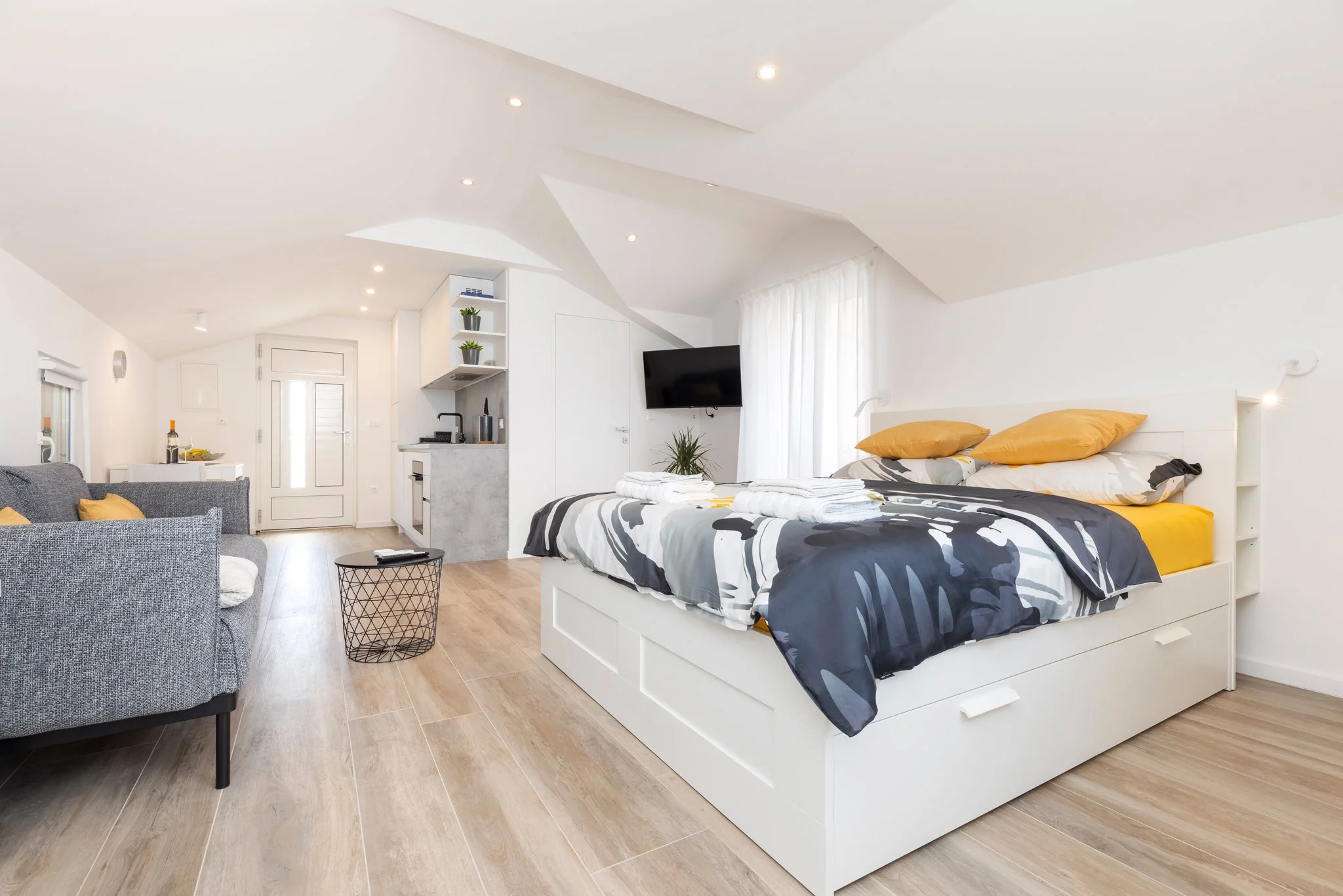 A cozy, modern studio apartment with a white bed, yellow pillows, black-and-white bedding, a flat-screen TV on the wall, a small kitchen area, and a sitting area with gray upholstered chairs. The room has white walls, a vaulted ceiling with recessed 