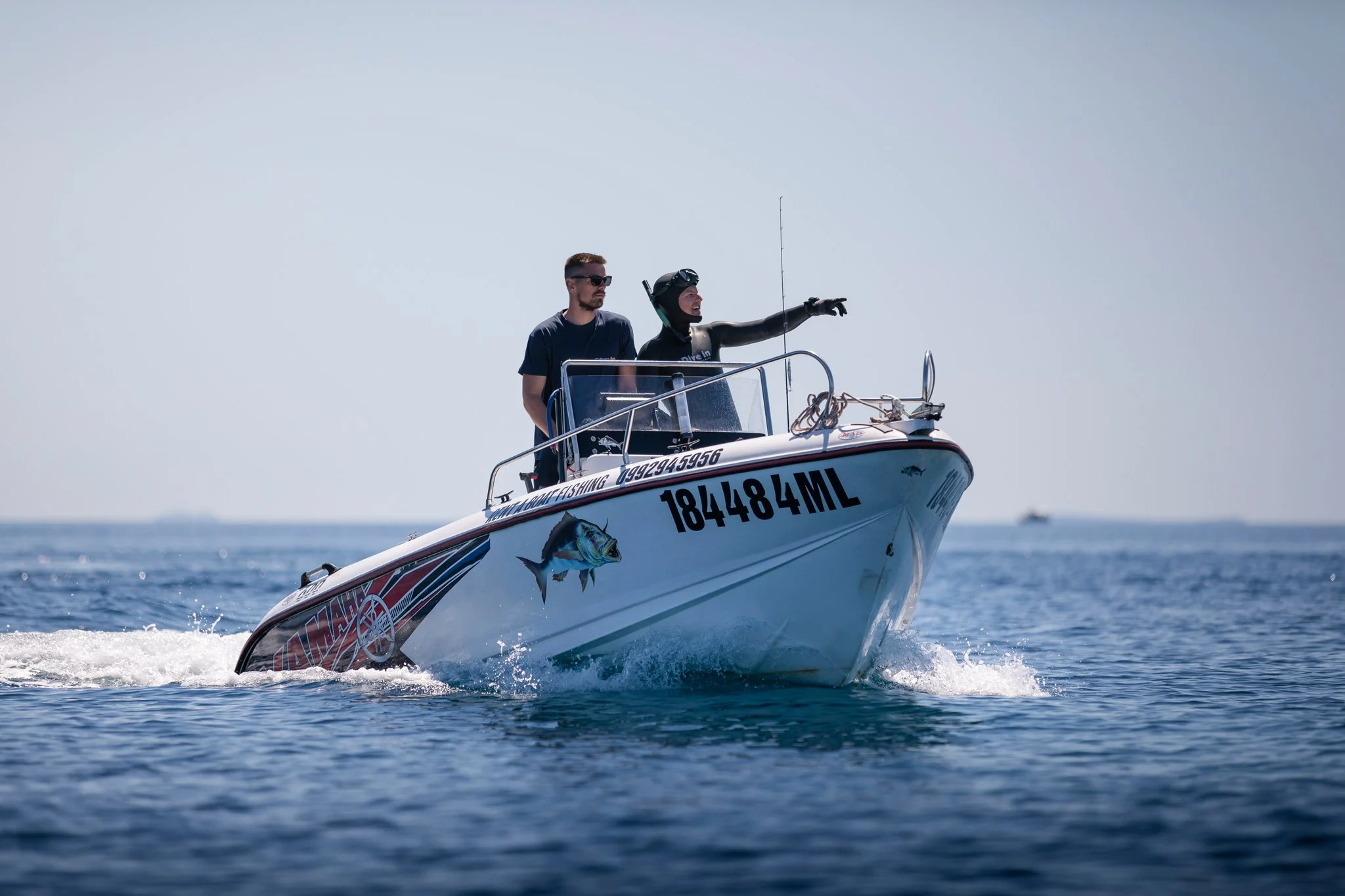 Two men on a speedboat on the water; one wearing sunglasses and the other wearing a wetsuit and gloves, pointing into the distance.
