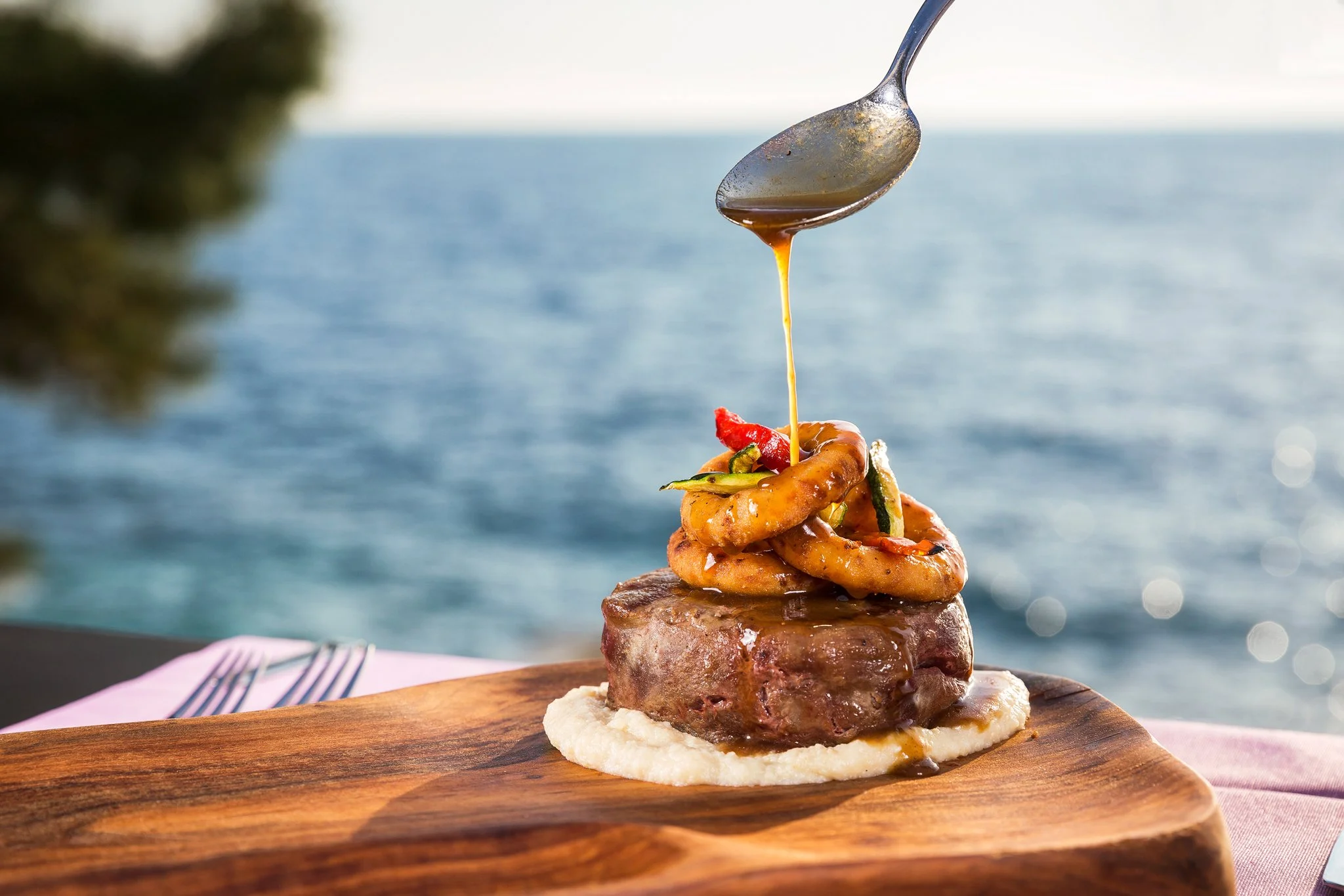 A close-up of a tall stack of fried onion rings with sauce and garnishes on top, served on a wooden board with a blurred ocean view in the background.
