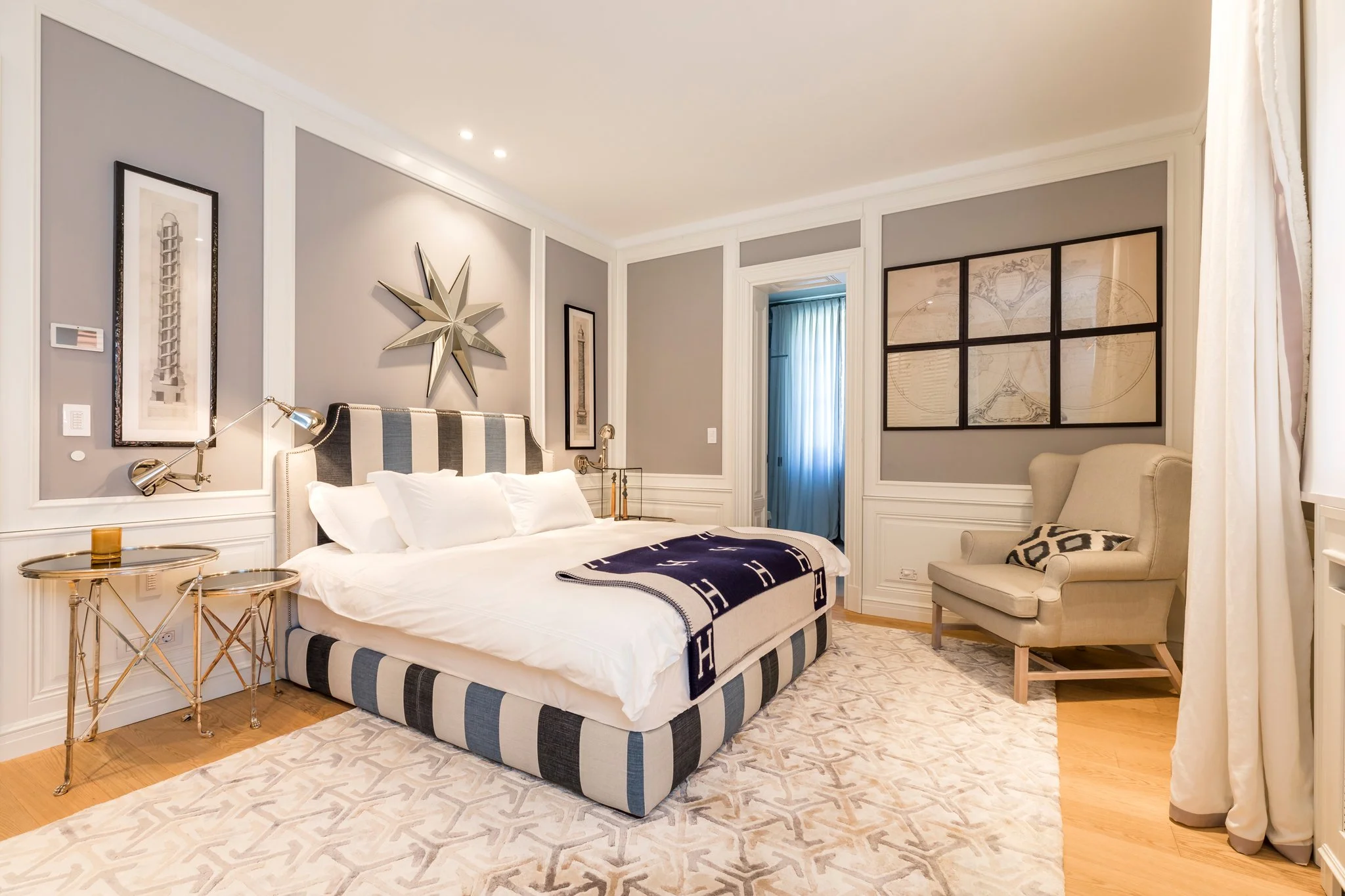 A stylish bedroom with gray walls, decorated with framed artwork, a star-shaped mirror, and a large bed with a striped upholstered headboard and navy throw. There are side tables with gold lamps, a beige armchair, and a window with white curtains. Th