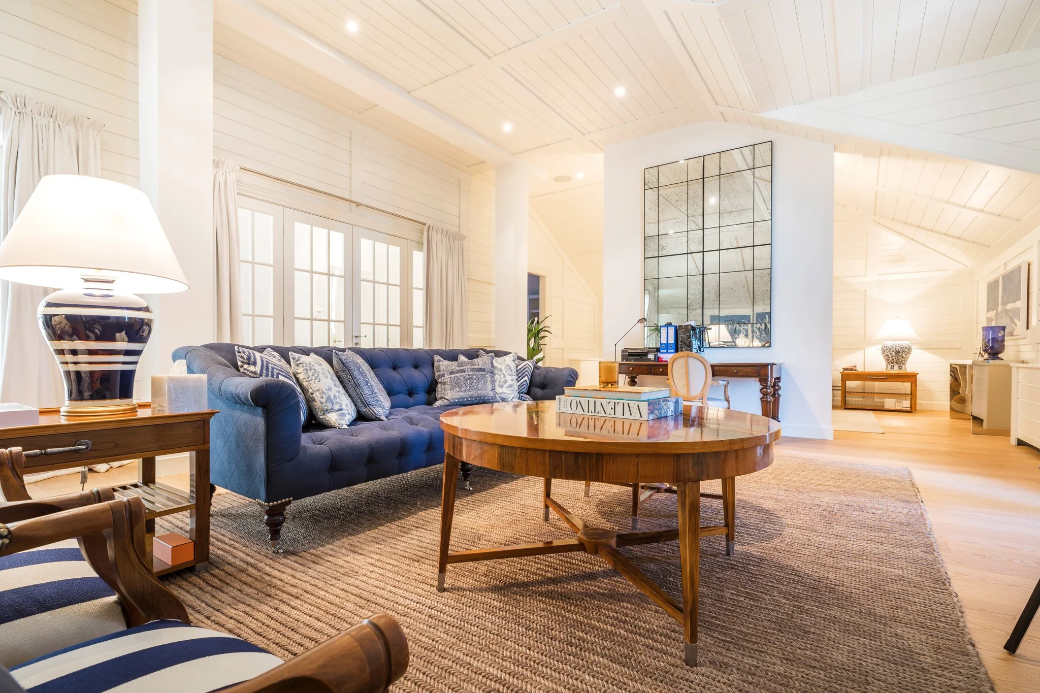 Bright living room with white wood-paneled ceiling and walls, featuring a dark blue tufted sofa with patterned pillows, a round wooden coffee table, a side table with a black and white striped lamp, large windows with white curtains, and framed artwo