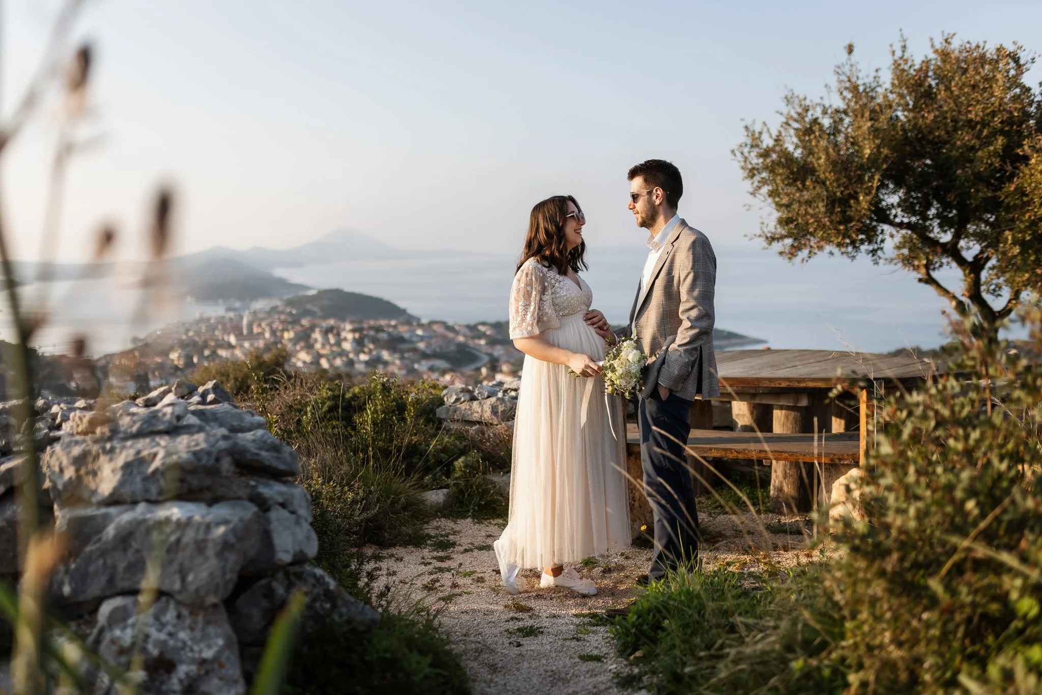 A couple dressed for a wedding, standing outdoors on a hilltop with scenic ocean and city views, holding hands and smiling at each other during sunset.