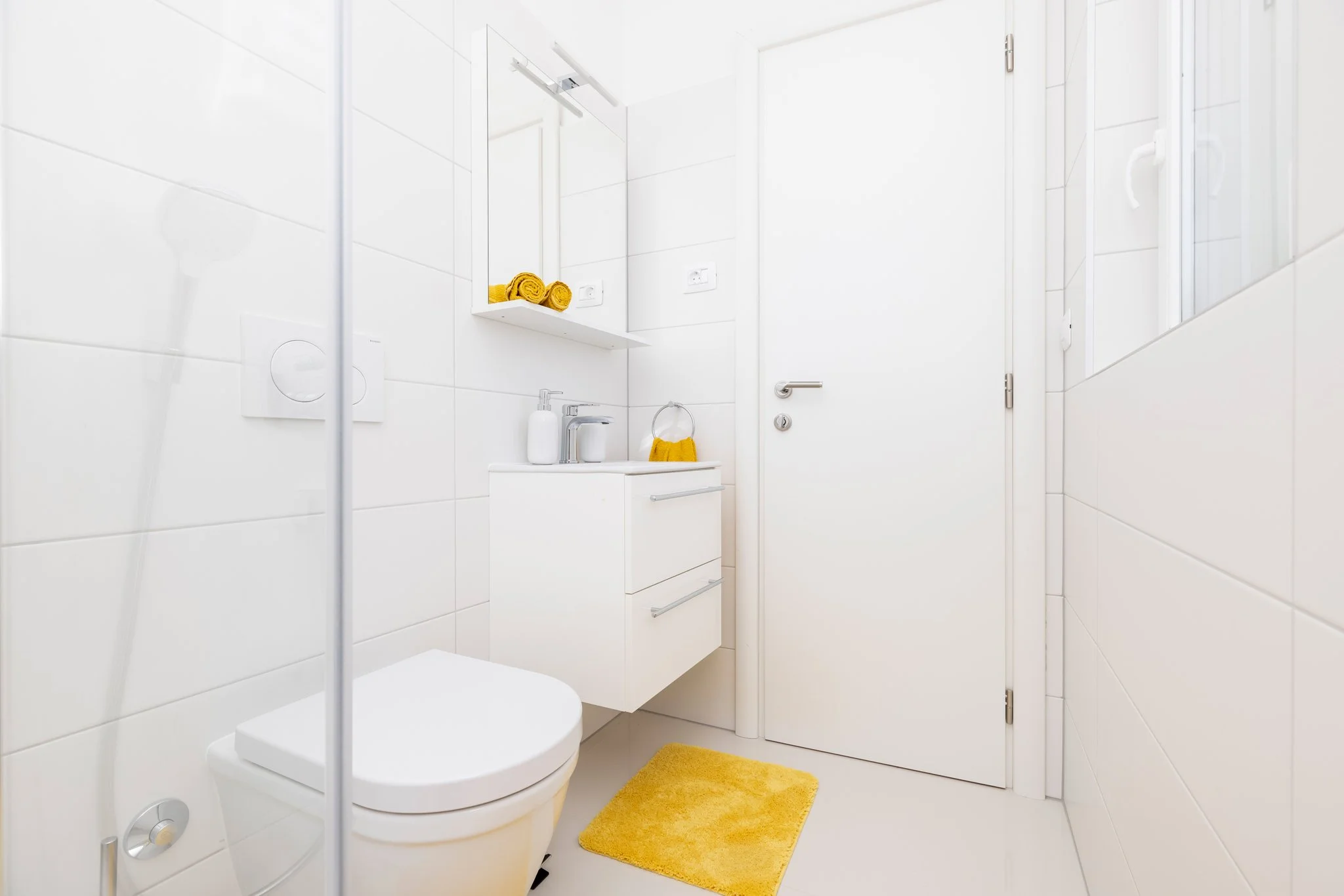 Small, modern bathroom with white tiled walls, a white toilet, and a white vanity with a small sink. There are yellow towels placed on a shelf and hanging from a ring, with matching yellow bath mats on the floor. A mirror is mounted above the vanity,