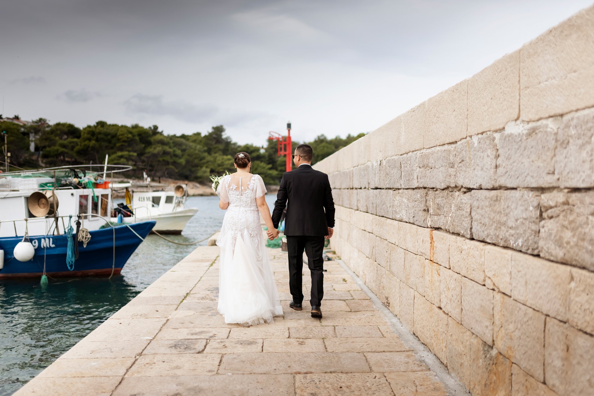 A bride and groom walking hand in hand along a harbor walkway, with boats docked on one side and a stone wall on the other, under cloudy skies.