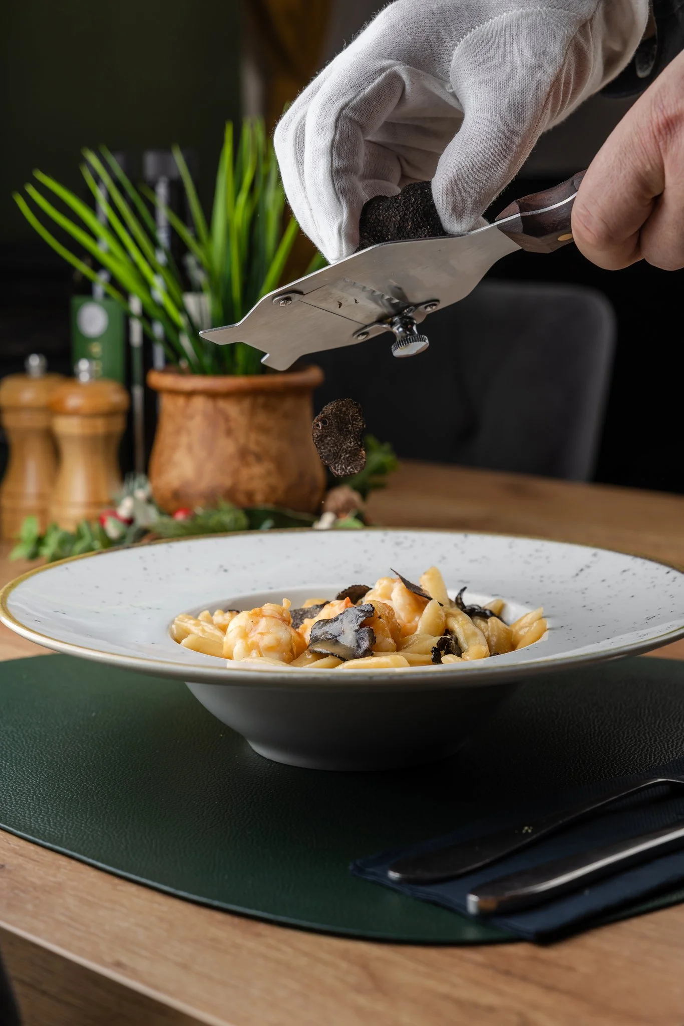 Close-up of a hand wearing a white glove slicing black truffle over a plate of pasta. In the background, there are kitchen items and a plant.