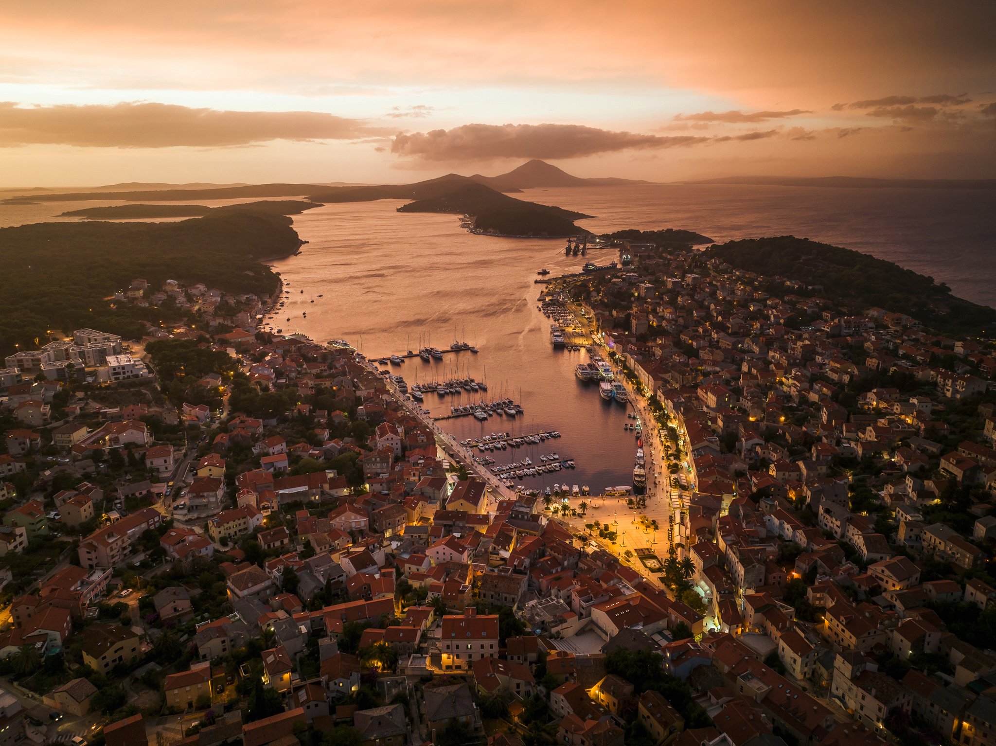 An aerial view of a coastal town at sunset, with houses, a marina with boats, and surrounding green hills.