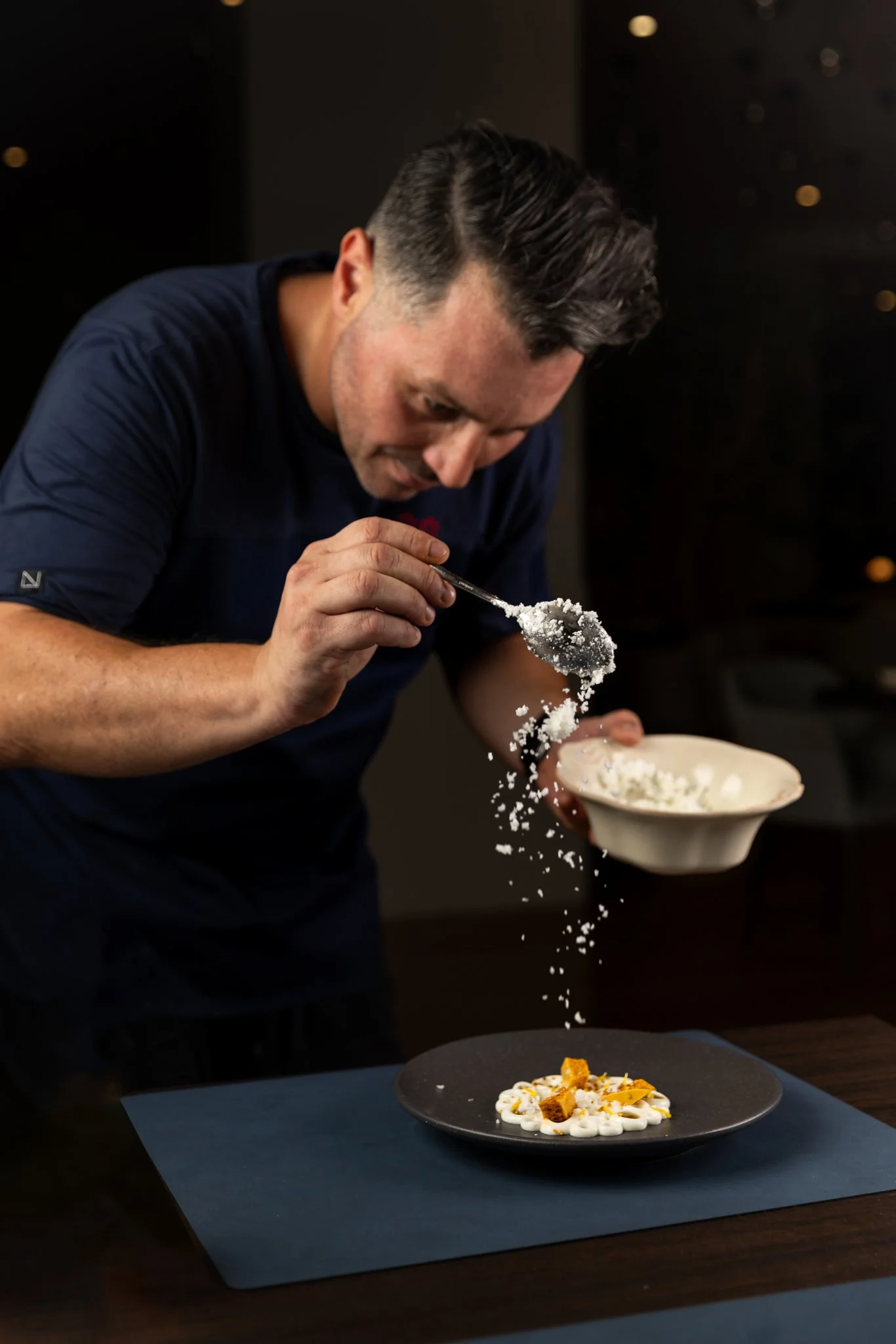 A chef garnishing a plated dish with salt or cheese in a professional kitchen setting.