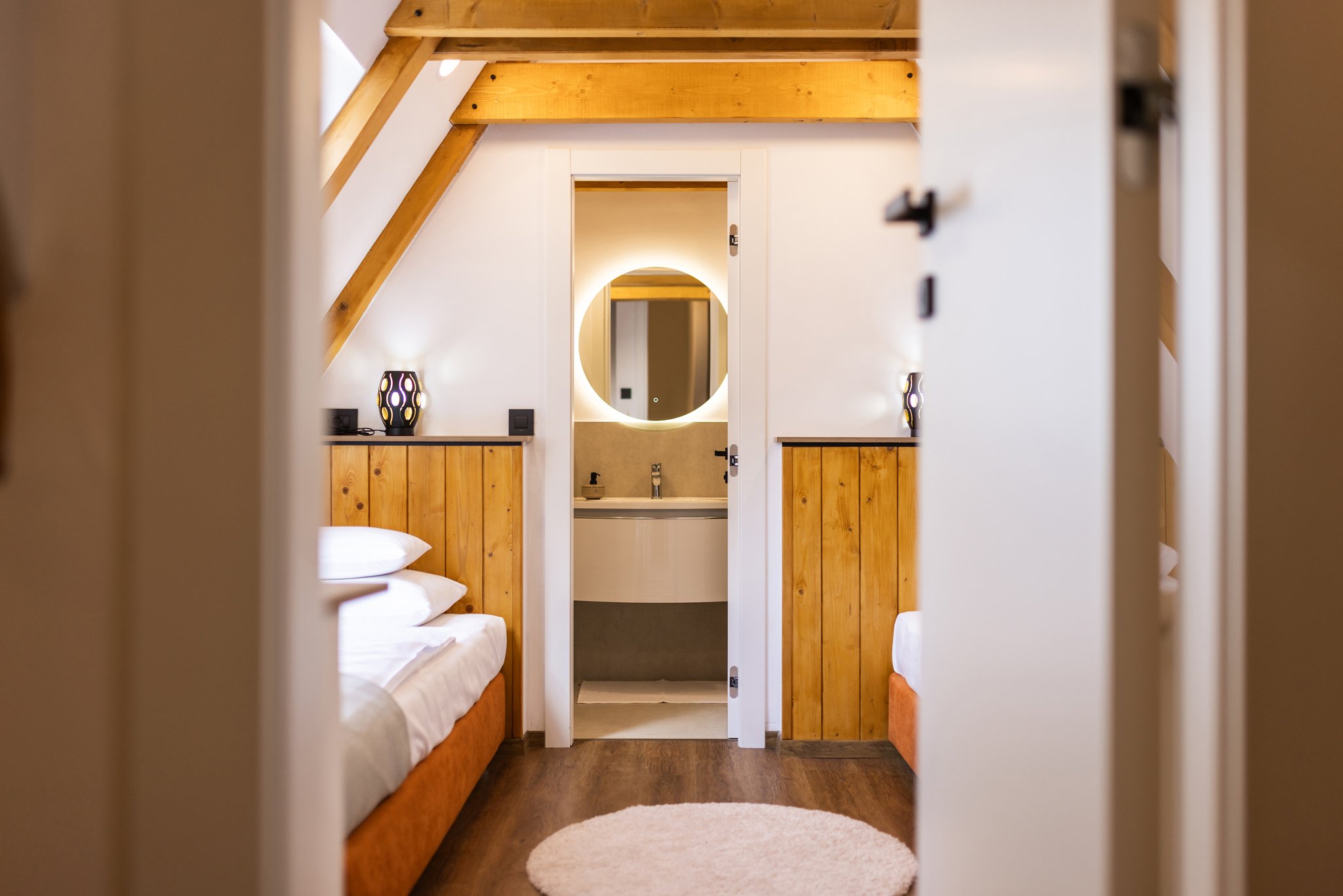 A cozy bedroom viewed through a partially open door, featuring two beds with white linens and wooden headboards, small bedside lamps, and a round beige rug on a dark hardwood floor. There is a small sink and mirror visible through an open doorway in 