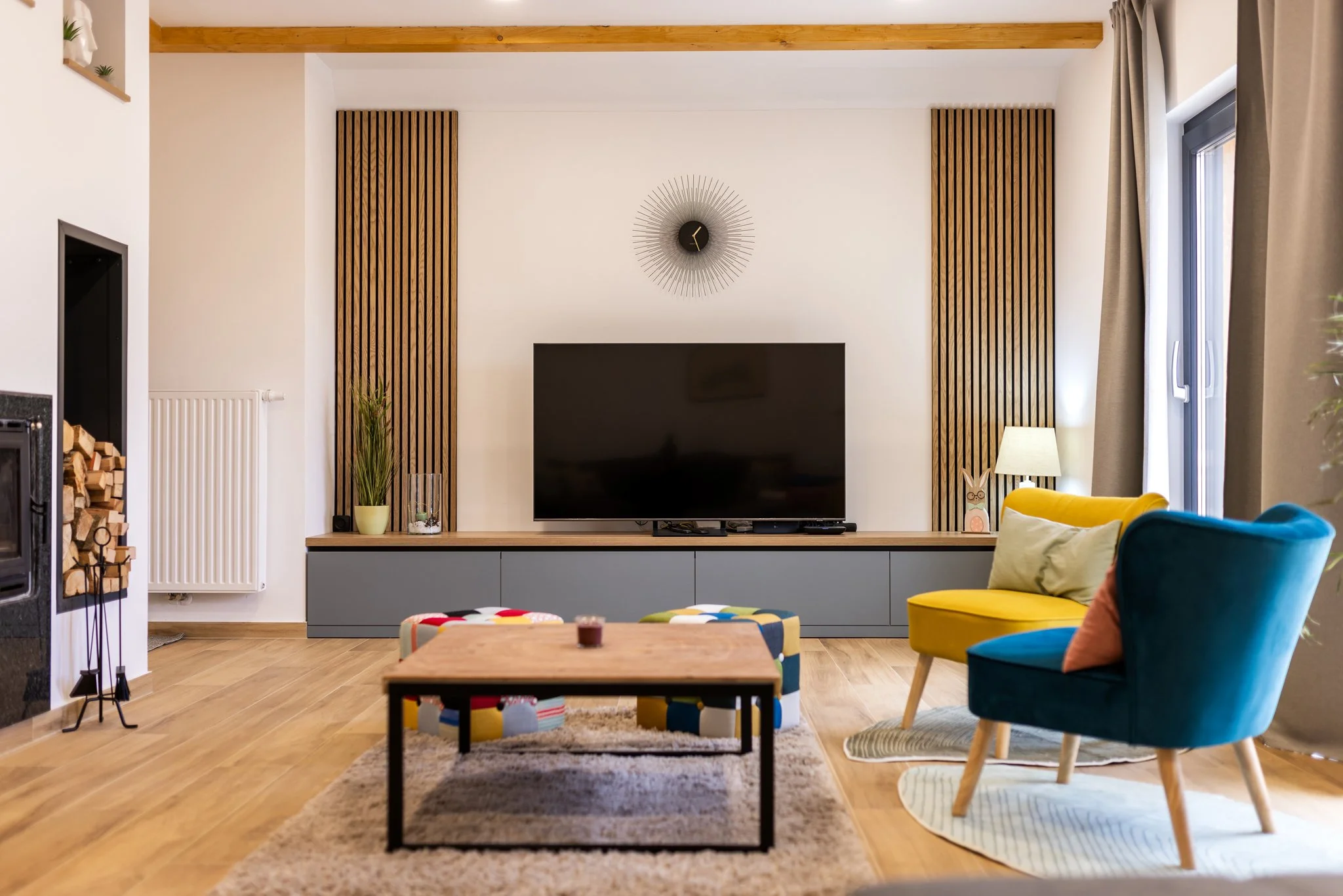 Living room with a modern interior, featuring a wooden accent wall, a flat-screen TV, a decorative wall clock, and colorful furniture including a yellow and teal armchair and a patterned ottoman. There are curtains on a window to the right, a lamp on