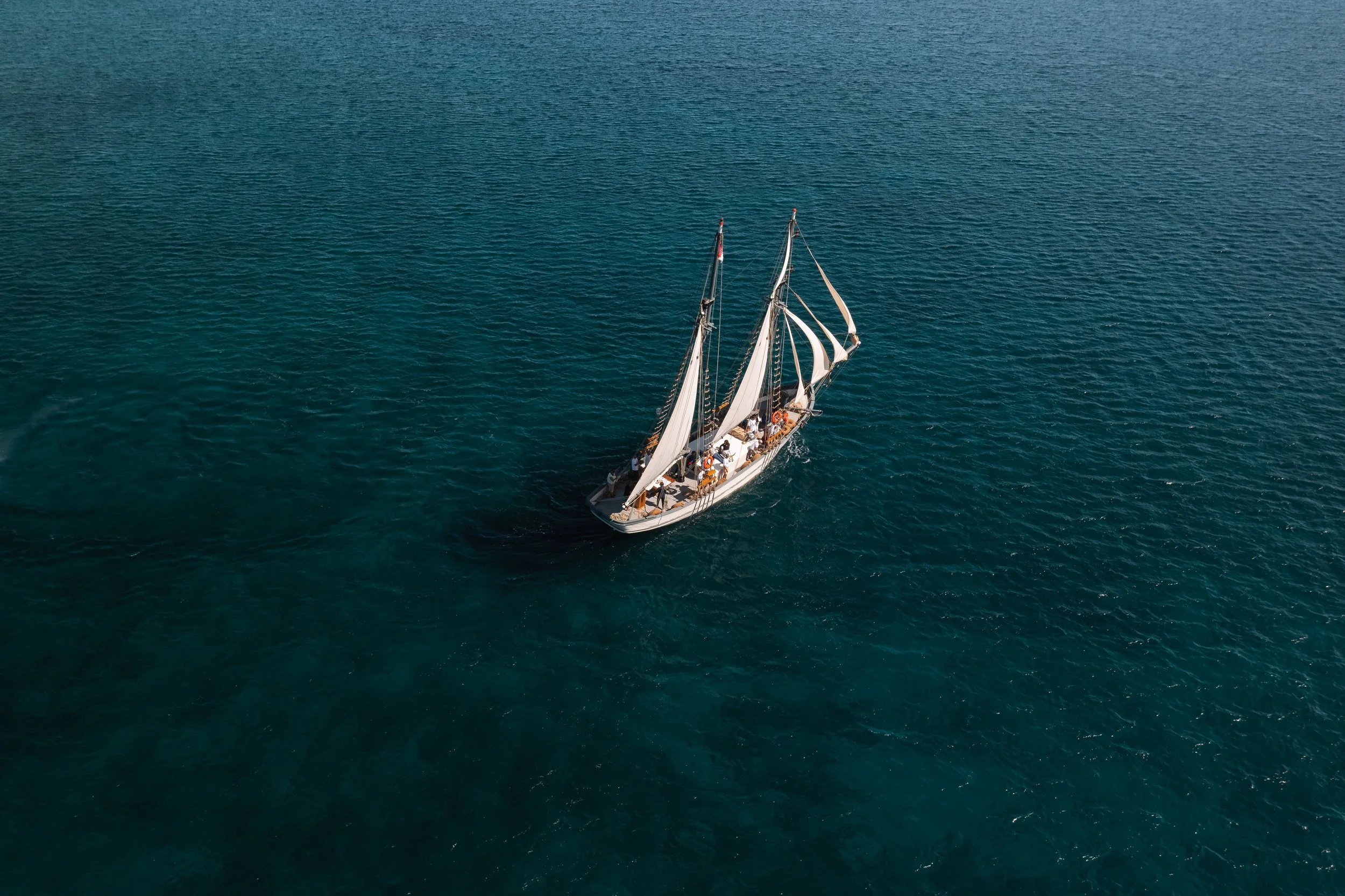 A sailboat with multiple sails sailing on a large body of water, viewed from above.