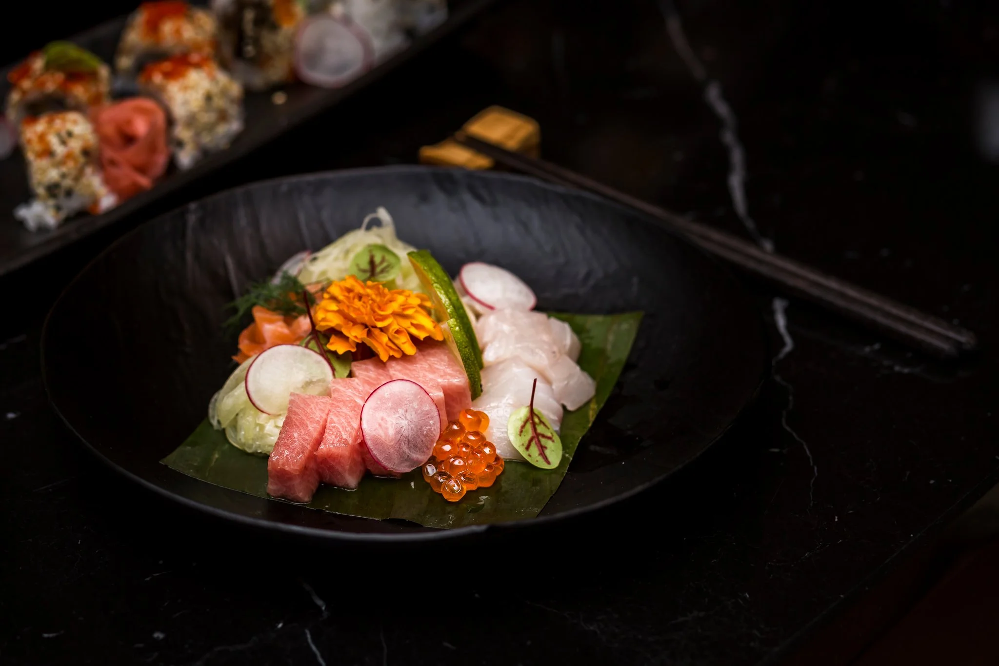 Sashimi platter with slices of raw fish, radish slices, cucumber, salmon roe, edible flower, and garnishes, on a black plate with a dark background.