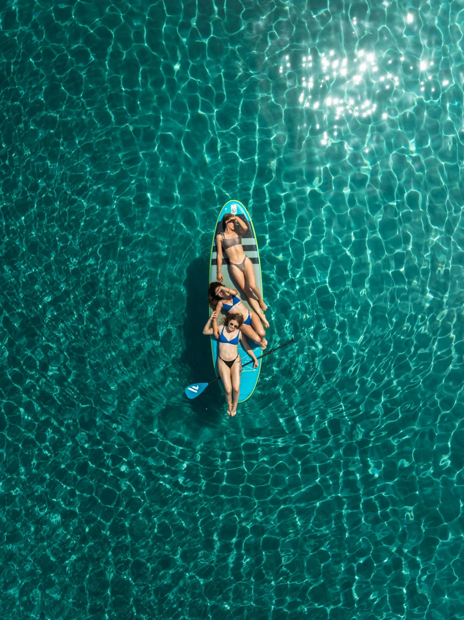 Top-down view of a woman in a bikini relaxing on a paddleboard in clear blue-green water. She is shielding her eyes from the sun with her hand, and her sunglasses are visible.