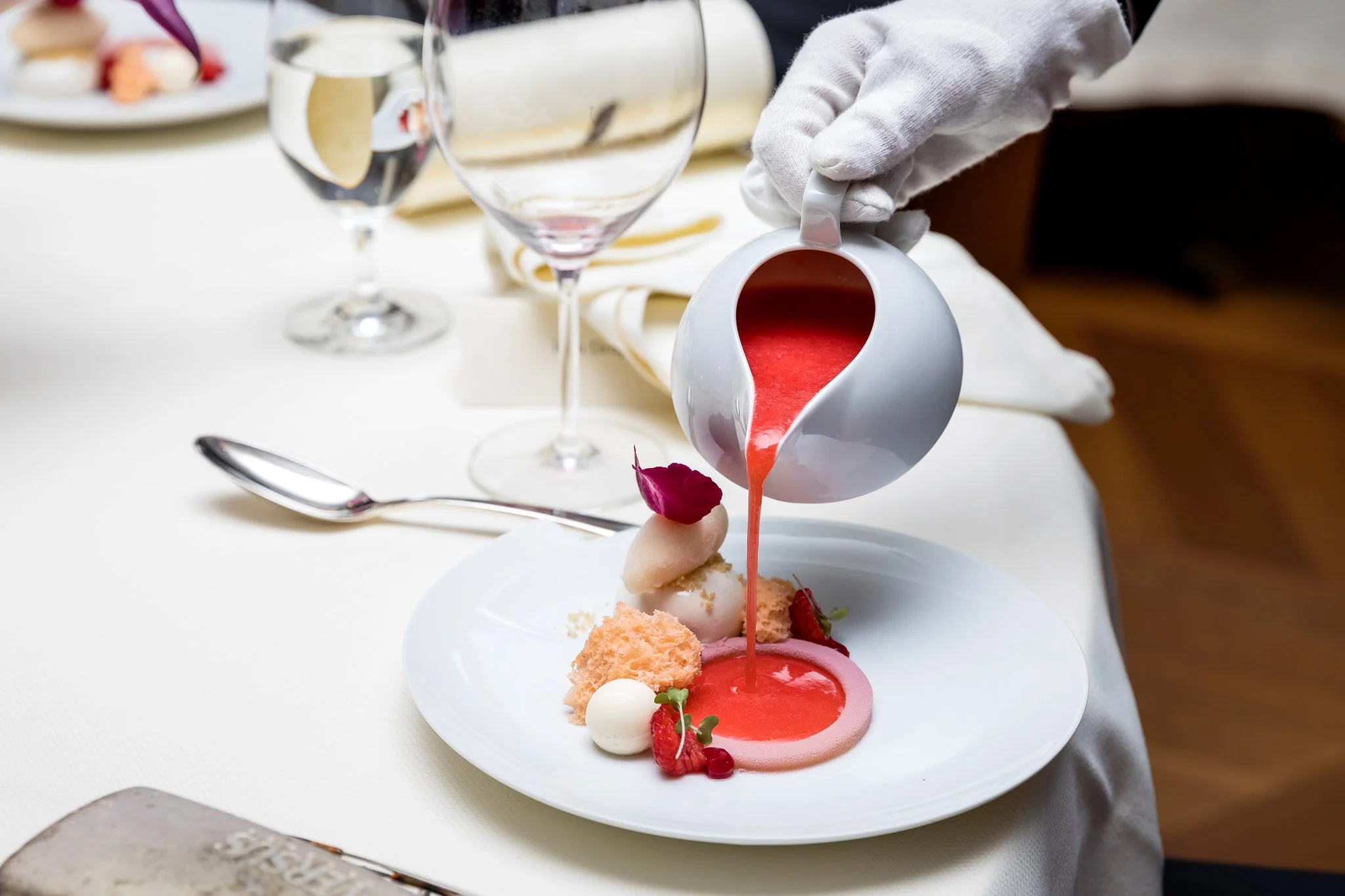 A chef pouring red sauce onto a plated dessert with various garnishes, on a white tablecloth, in a fine dining setting.