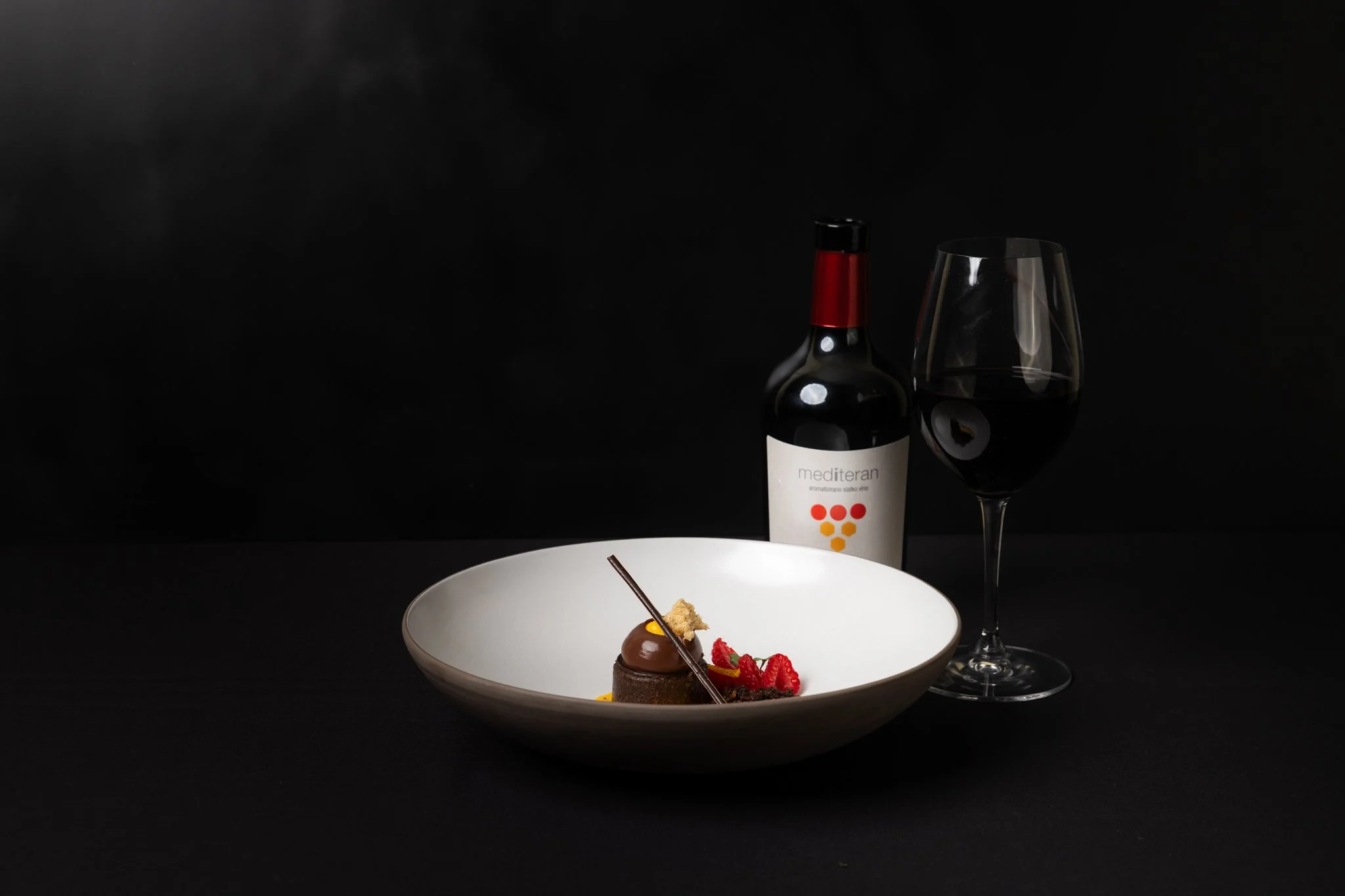 A white bowl with dessert topped with chocolate, strawberries, and a decorative stick. Behind it is a wine bottle and a glass of red wine on a black background.