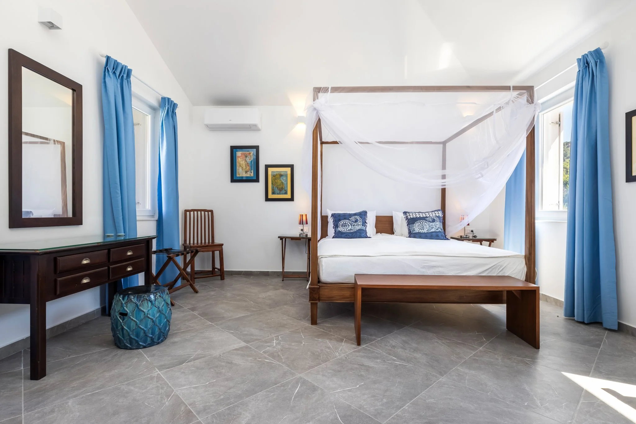 A bedroom with a wooden four-poster bed with white canopy and blue checkered pillows, blue curtains on windows, framed artwork on white walls, gray tiled floor, wooden chairs, and a dark dresser.