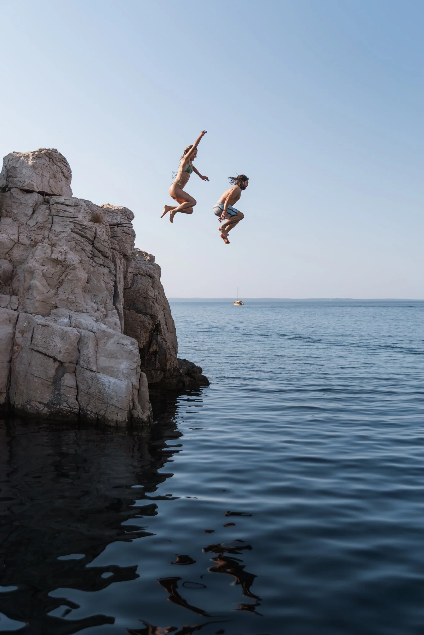 Two people jumping off a rocky cliff into the water during daytime, with a boat in the distant water and a clear blue sky.