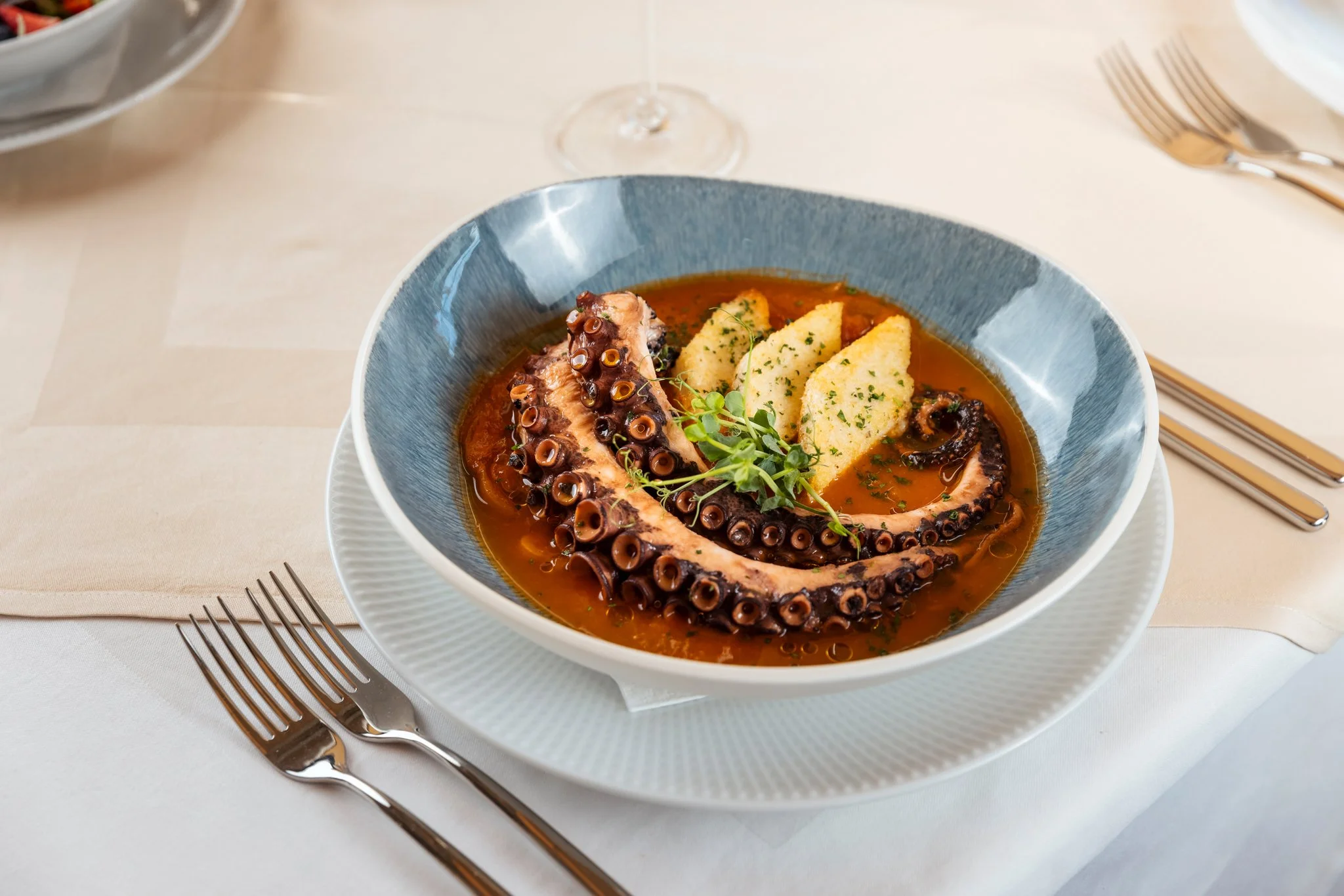 Bowl of grilled octopus with potatoes and herbs, garnished with microgreens, on a white table set with forks and a napkin.