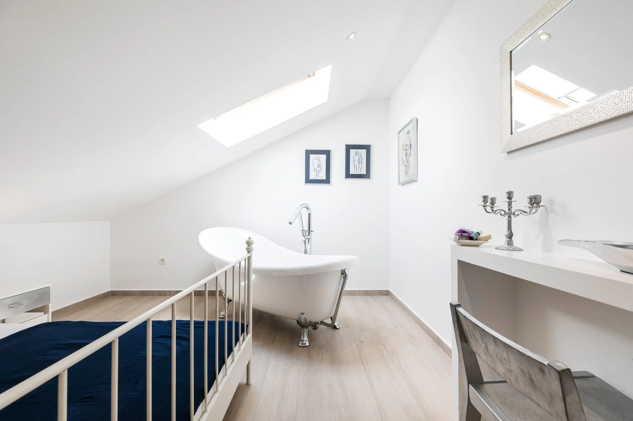 A bright, white bathroom with skylights, a clawfoot tub, framed line art on the wall, a silver candelabrum, and a dark blue bedspread partially visible in the foreground.