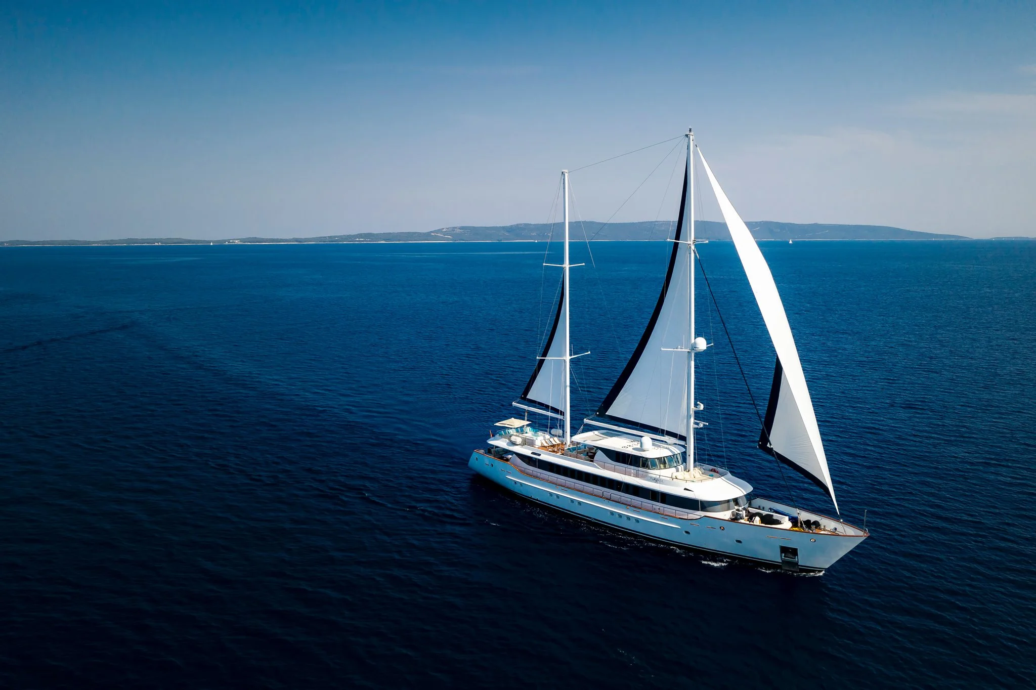 A large white sailing yacht with black and white sails is on calm blue water, with a distant landmass on the horizon under a clear sky.