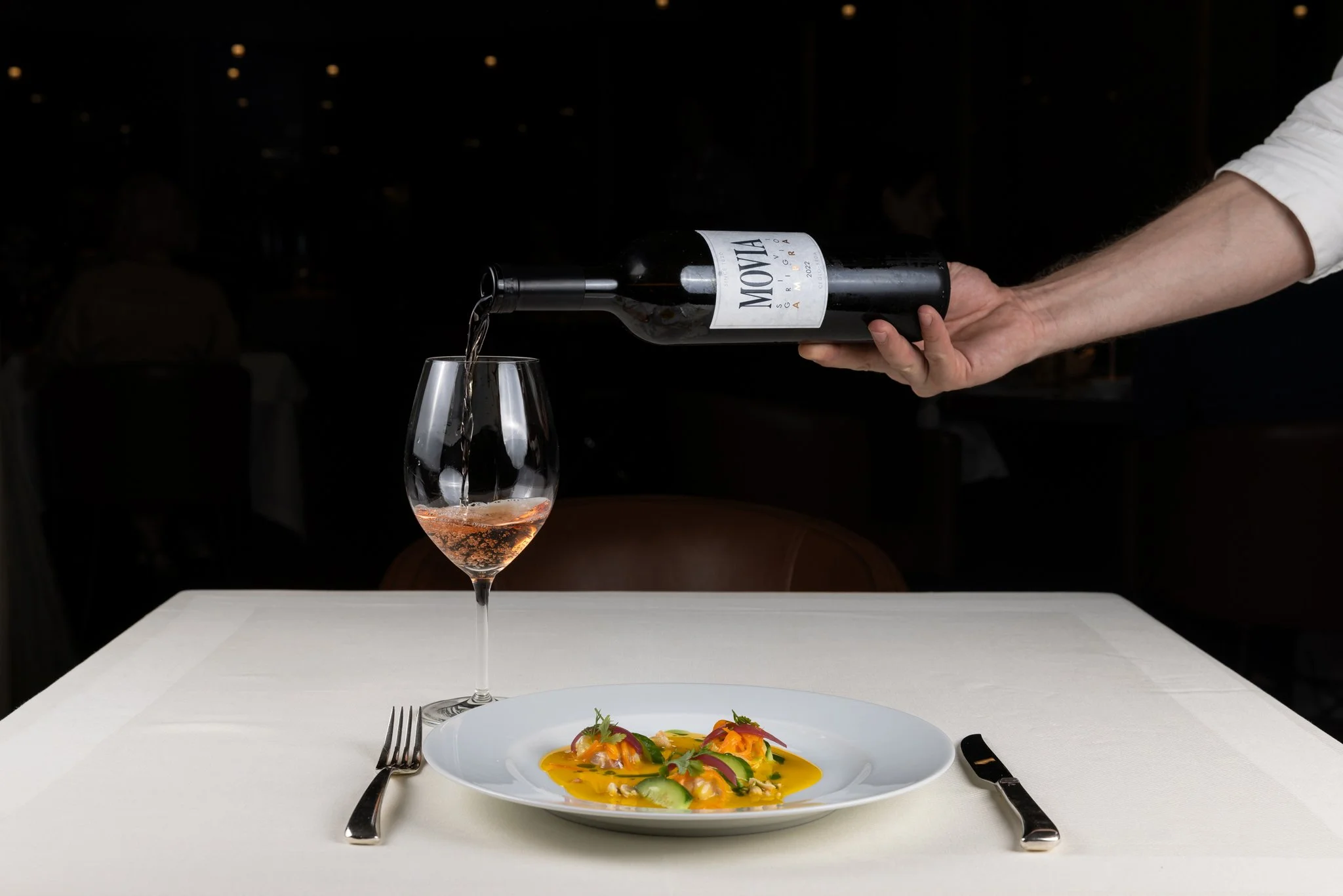 A restaurant scene with a glass of rosé wine being poured from a bottle labeled "Movia" into a wine glass. In the foreground, there is a white plate with an artistically plated dish garnished with herbs, and a fork and knife placed on each side of th