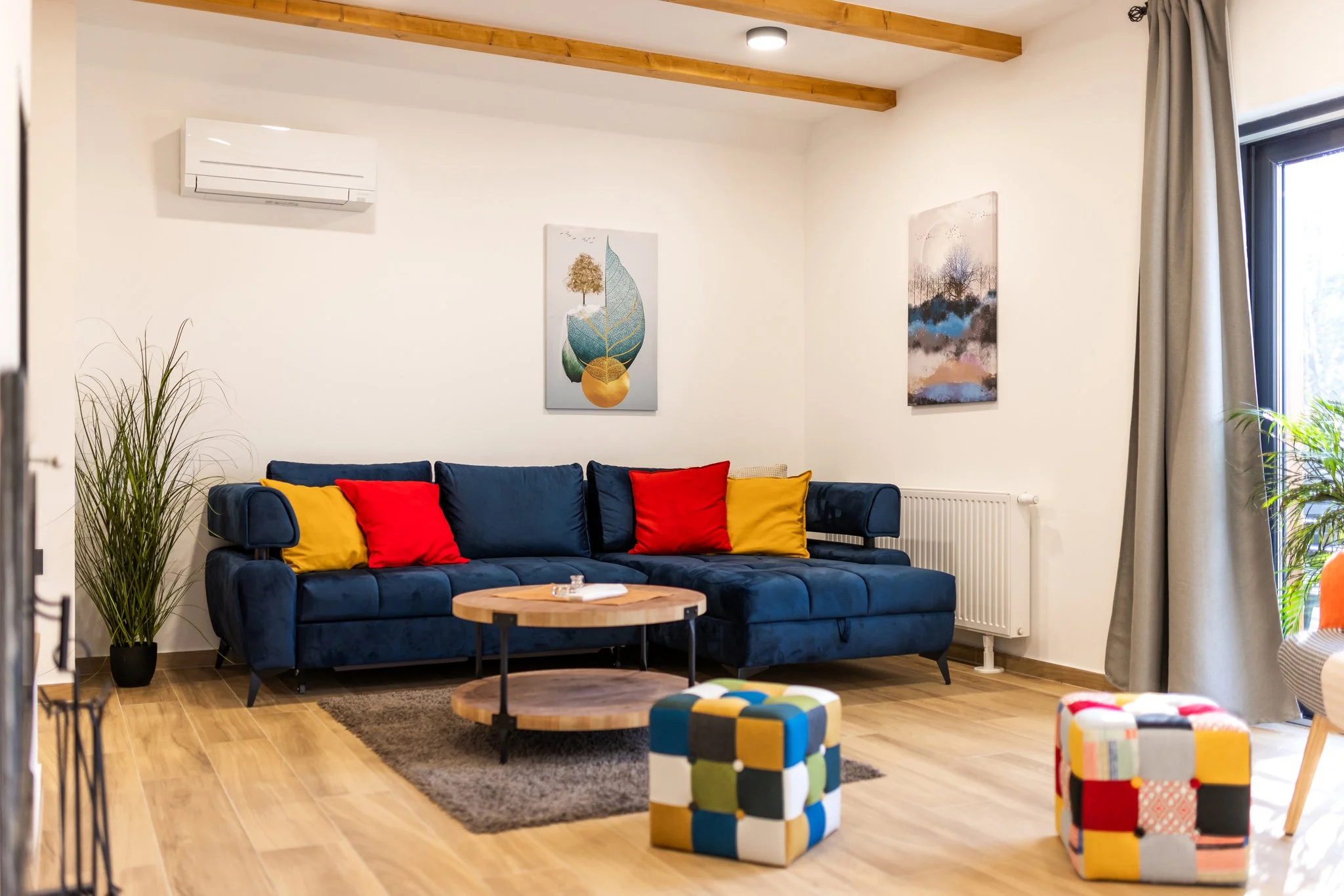 Living room with wooden floors, beige walls, a blue sofa with red and yellow pillows, artwork on the wall, a small round coffee table, plants, and four multicolored cube stools.