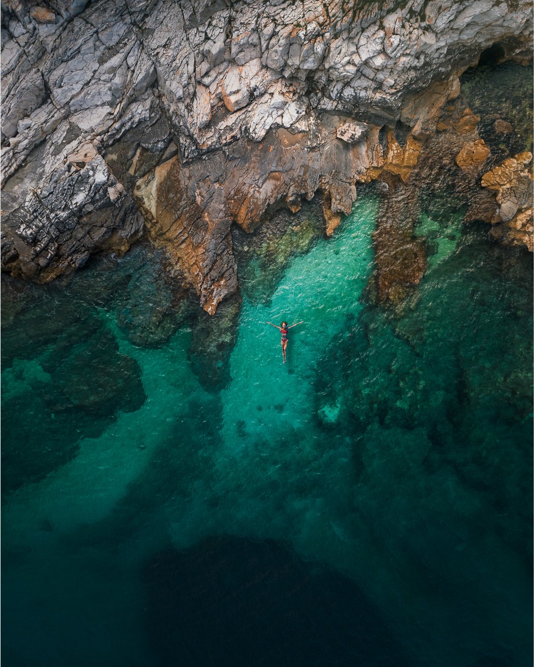 A person in a red swimsuit is floating in turquoise water near a rocky cliffside.