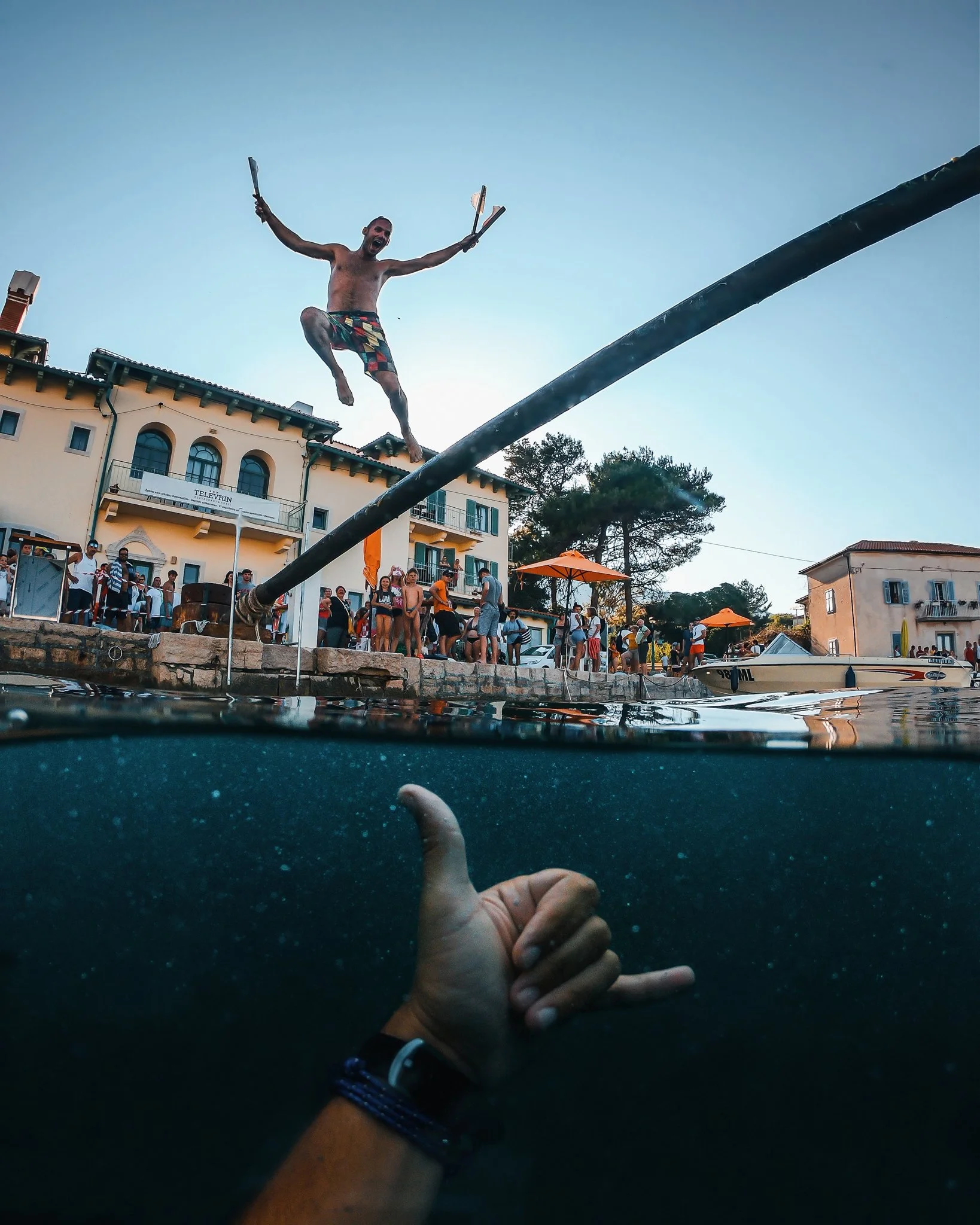 A person swimming and giving a thumbs-up in a pool, with a view from underwater looking up at another person jumping into the pool, holding a phone in one hand and making a gesture with the other, with spectators and buildings in the background durin