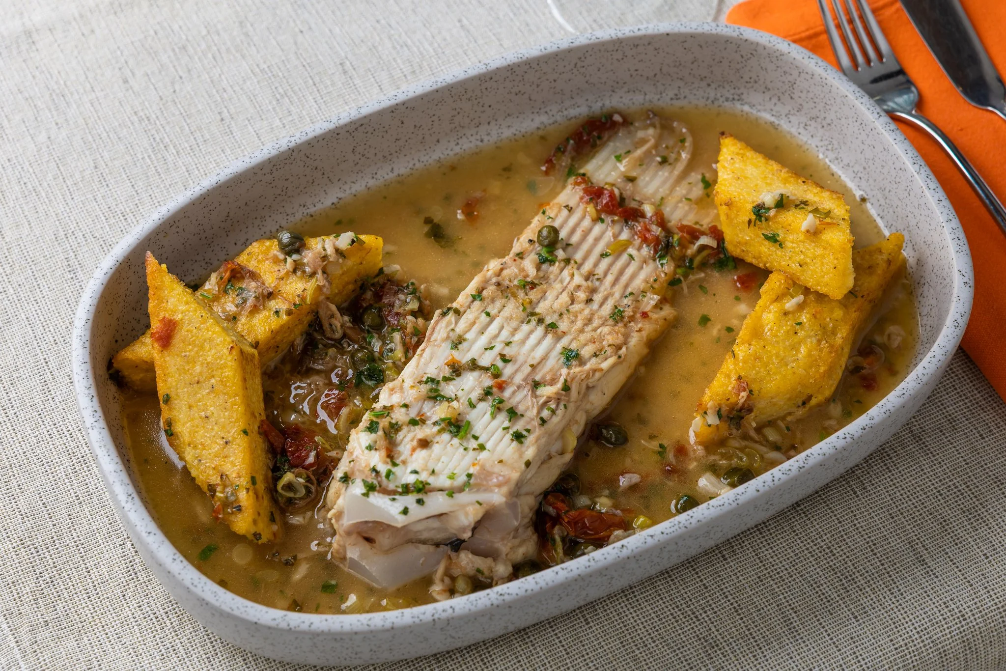 Baked fish fillet with herbs, served in a bowl with broth, garnished with chopped herbs, accompanied by fried polenta squares.