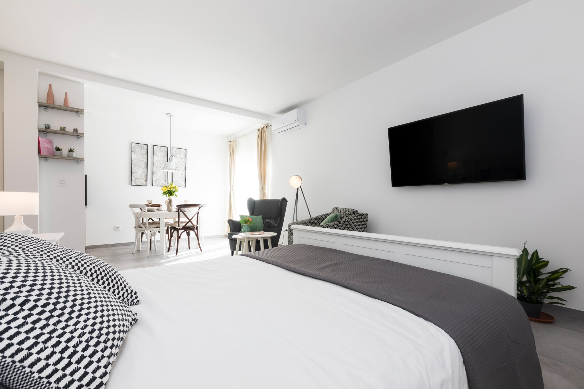 A modern, minimalistic open-plan living room with a bed in the foreground, a dining table with chairs, a black armchair, a wall-mounted TV, and decorative artworks on the wall, all in a bright and airy space with white walls and curtains.