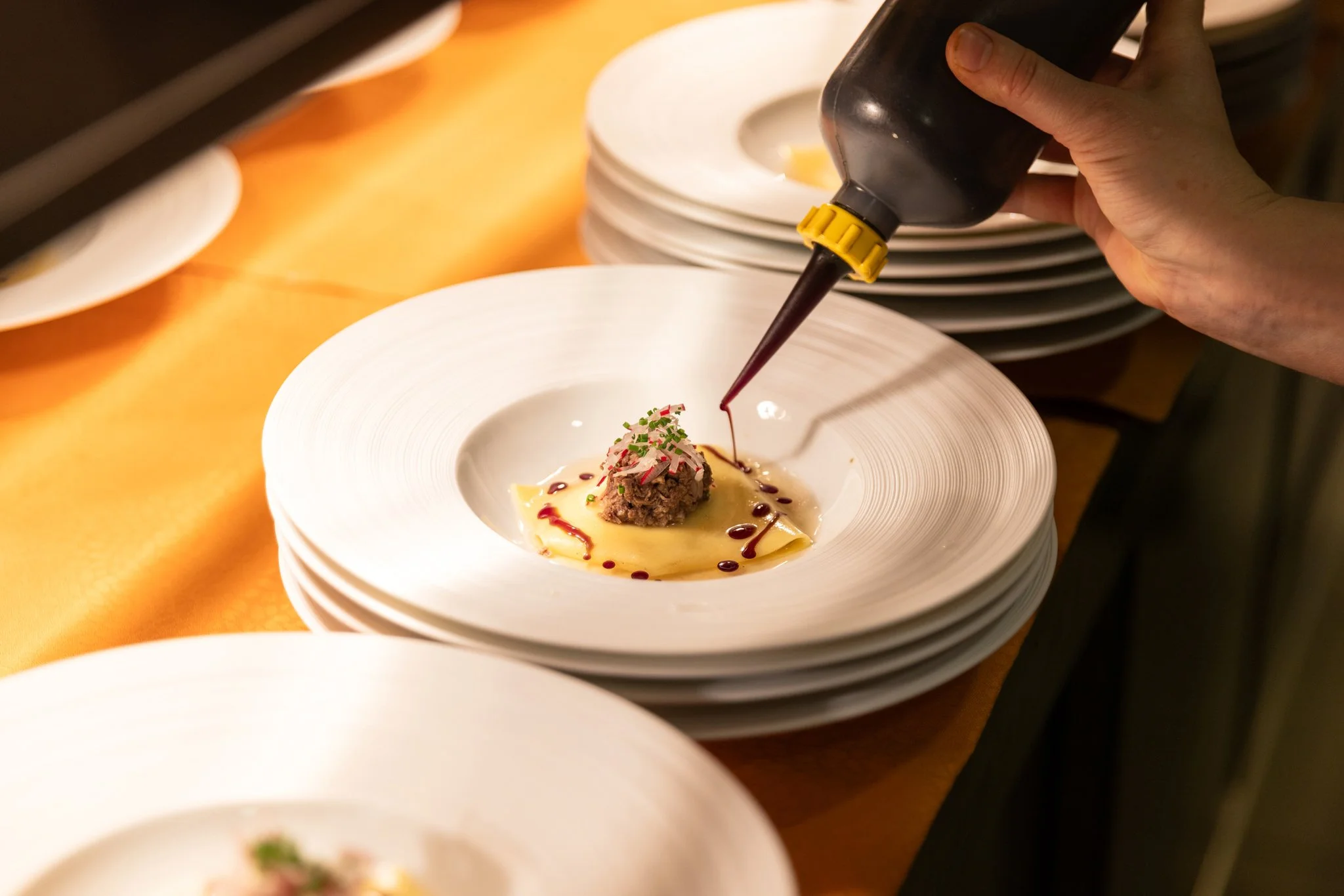 A hand holding a squeeze bottle drizzling sauce over a plated dish with diced meat, served on a white plate with a yellow sauce beneath and garnished with chopped herbs, set among the stacks of similar white plates on a wooden surface.