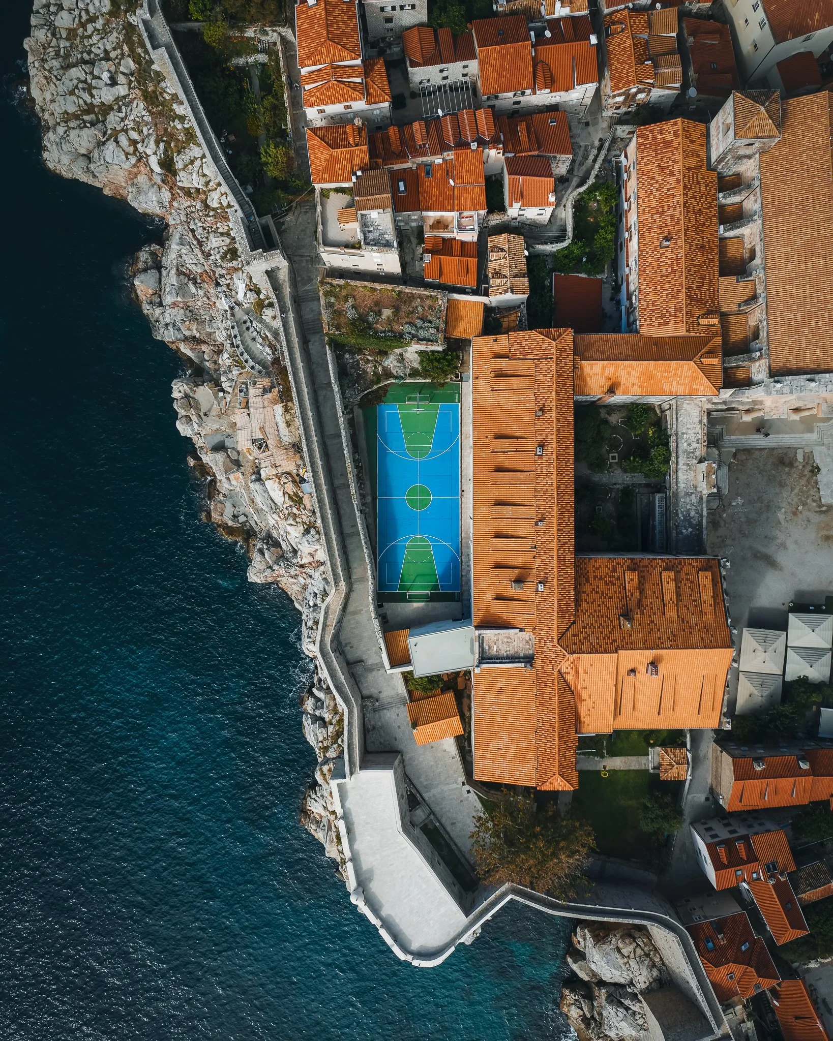 An aerial view of a coastal town with a fortress wall along the rocky shoreline. The town has buildings with orange-tiled roofs, and there's a colorful basketball court surrounded by the buildings. The deep blue sea is on the left side of the image.