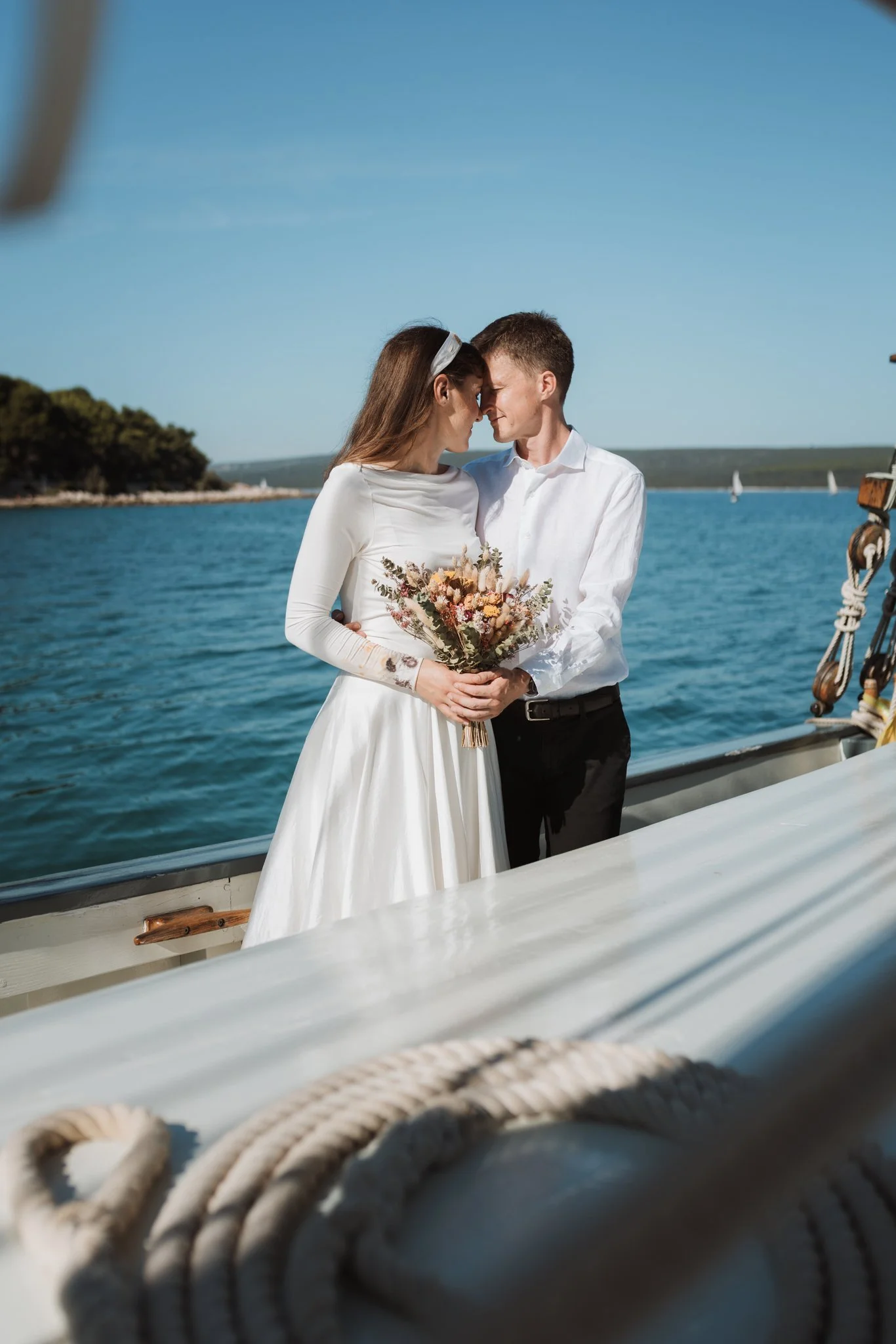 A couple dressed in wedding attire, standing close with foreheads touching, holding a bouquet on a boat near a body of water with sailboats in the background.