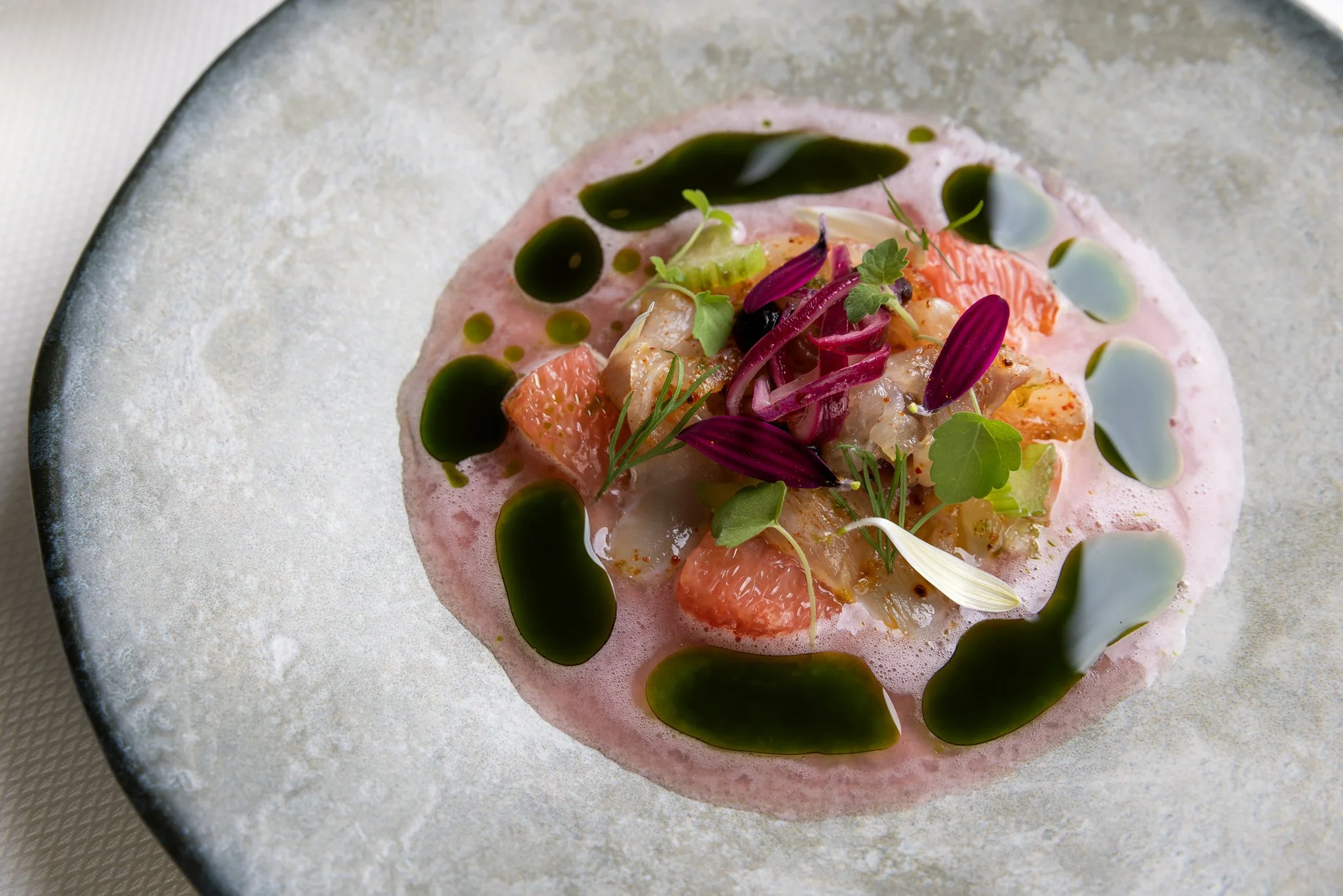 Close-up of a gourmet dish featuring grapefruit segments, microgreens, and drizzles of green and white sauces, served on a textured, off-white plate.