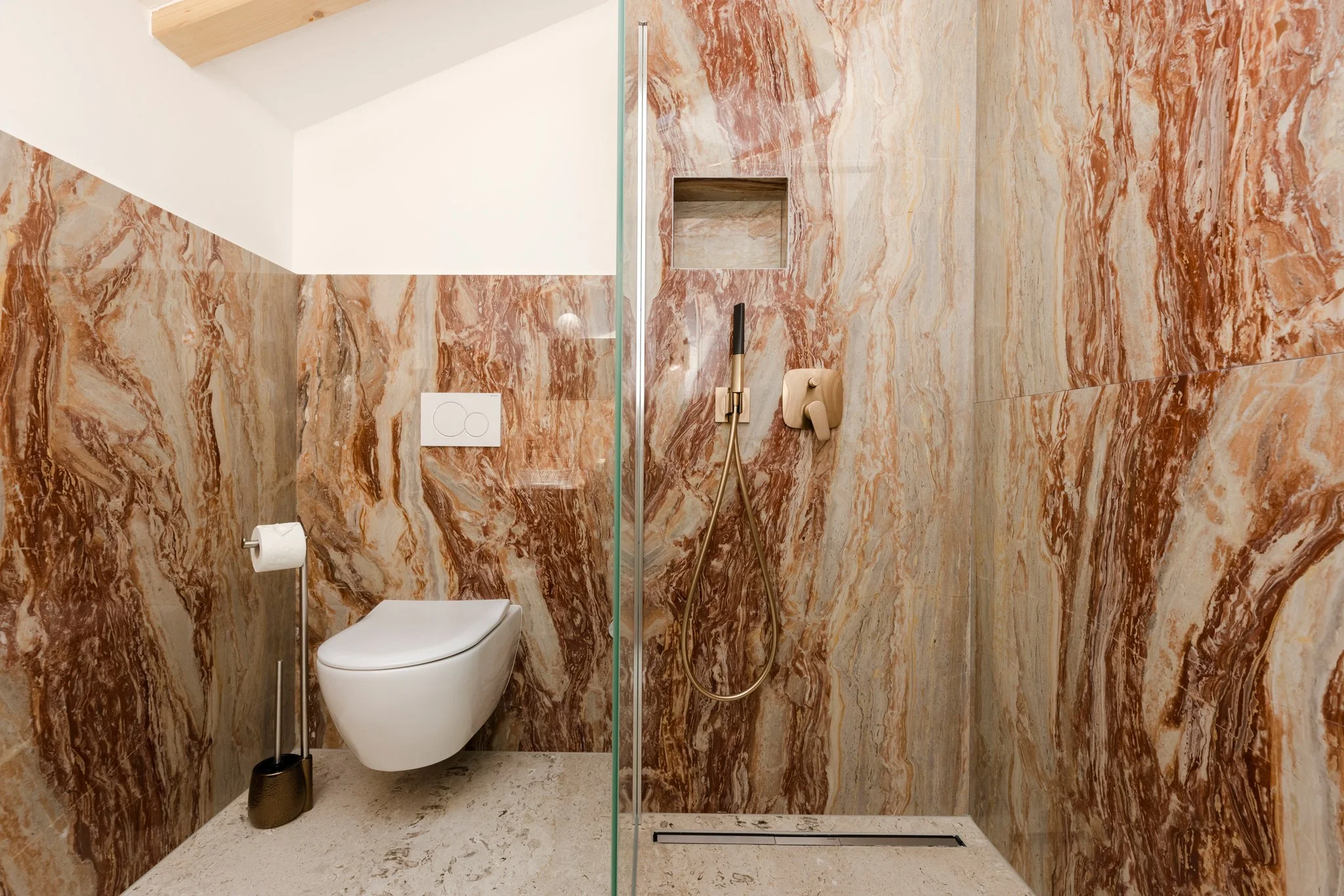 Bathroom with marble walls, a white toilet, a toilet paper roll, a black toilet brush, a shower area with a rainfall shower head and a handheld shower with gold fixtures, and a small built-in shelf in the wall.