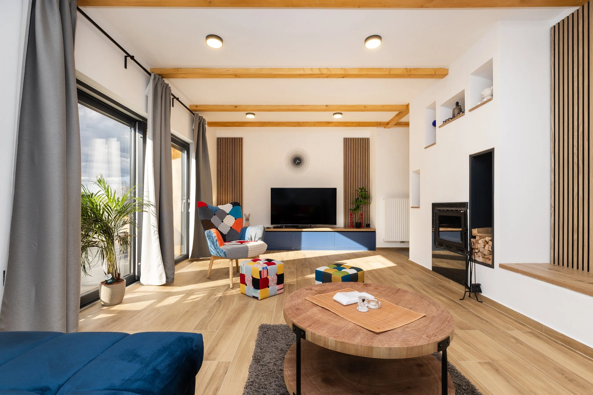 Modern living room with large windows, gray curtains, a blue sofa, colorful patterned armchair, wooden accents, flat-screen TV, fireplace, and decorative items on shelves.
