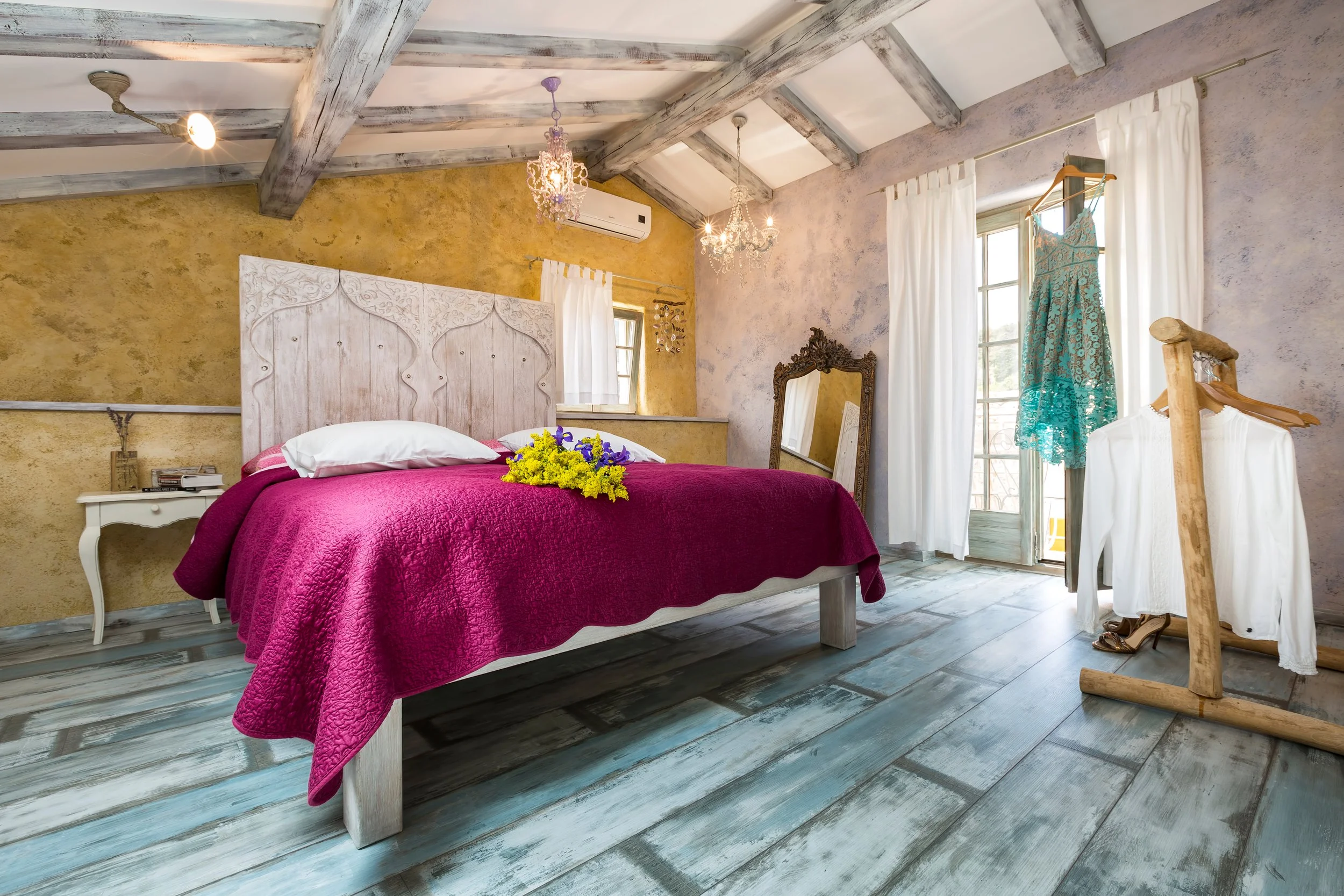 A bedroom with a rustic style, featuring a bed with a deep pink quilt, a floral arrangement on the bed, an ornate mirror, white curtains, and a dress hanging on a wooden stand. The ceiling has exposed wooden beams and two chandeliers hanging, while t