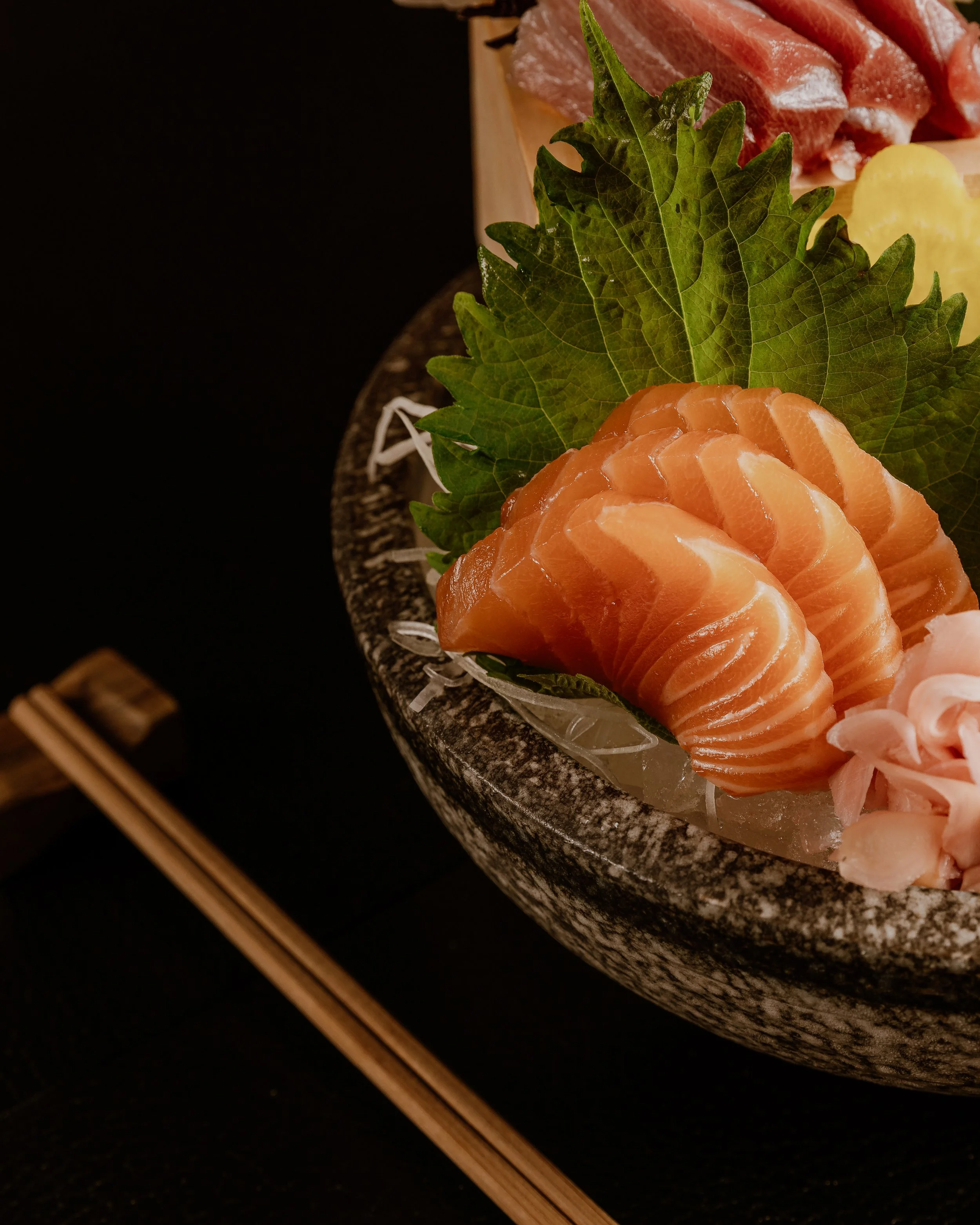 Close-up of sliced raw salmon sashimi on a plate garnished with green shiso leaves, with a dark background and chopsticks in the corner.